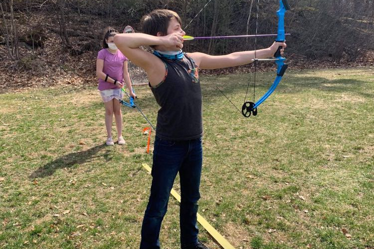 Image of Boynton students at archery club. A student in the background has their bow pointed toward the ground while they watch a student in the foreground who is about to shoot an arrow. 