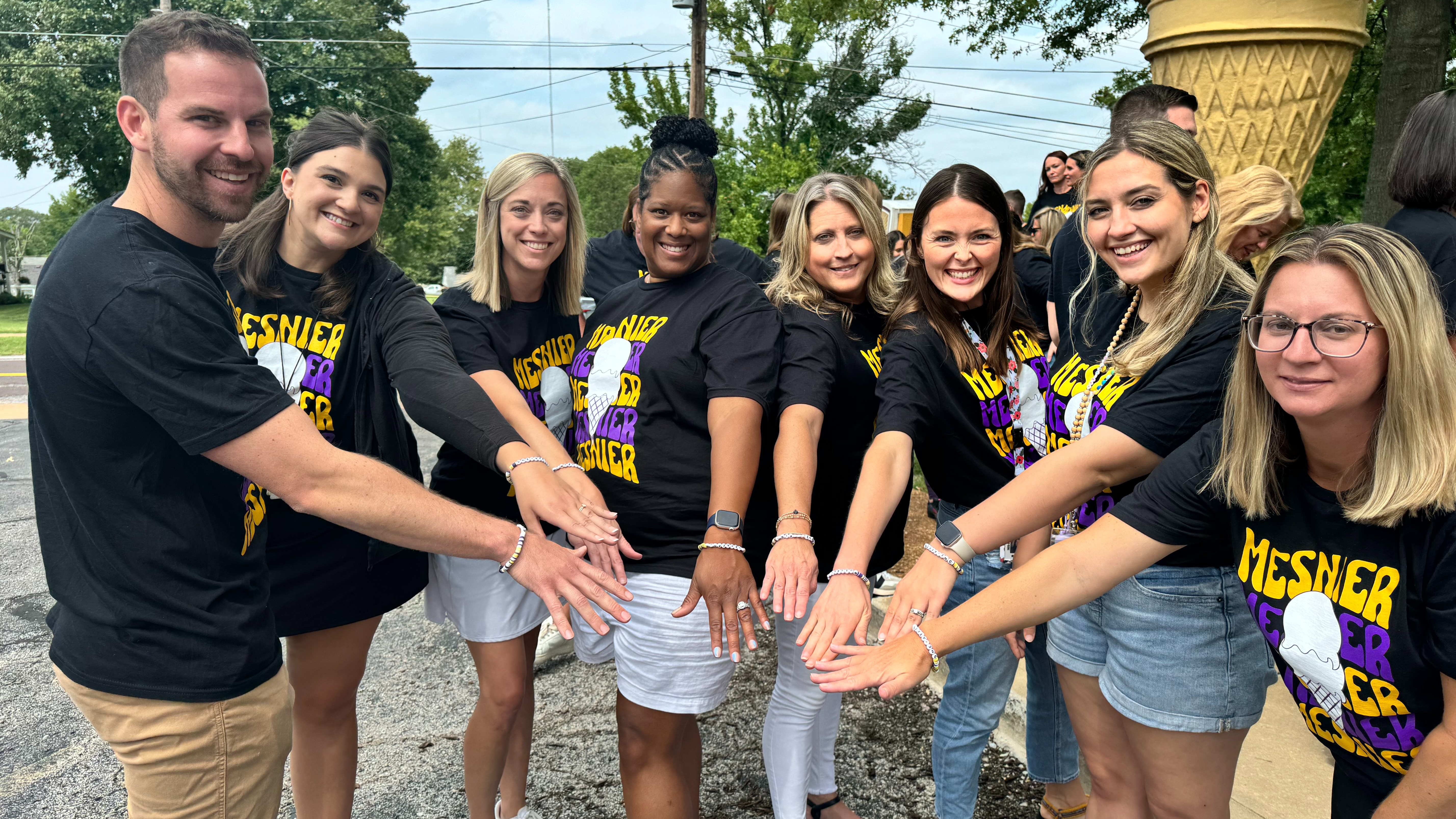 A group of eight people, consisting of men and women, stand together outdoors, smiling and extending their hands forward in unison. They are wearing matching black T-shirts with bold yellow and purple text that reads "MESNER" alongside an image of an ice cream cone. The background features a large, decorative ice cream cone structure and a crowd of people wearing similar shirts, indicating a community event or gathering.