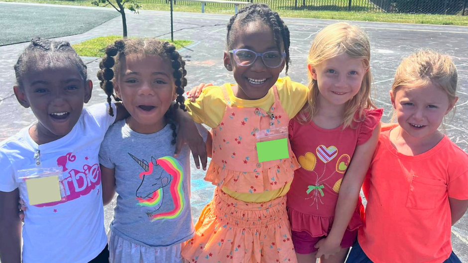 Five young girls stand together on a playground, smiling with their arms wrapped around each other. They are dressed in colorful outfits, including a "Barbie" T-shirt, a unicorn-themed dress, a bright orange ruffled set, a red top with heart designs, and a neon orange shirt with a denim skirt. The sun is shining, casting shadows on the chalk-covered pavement, while a fence and grassy area are visible in the background.