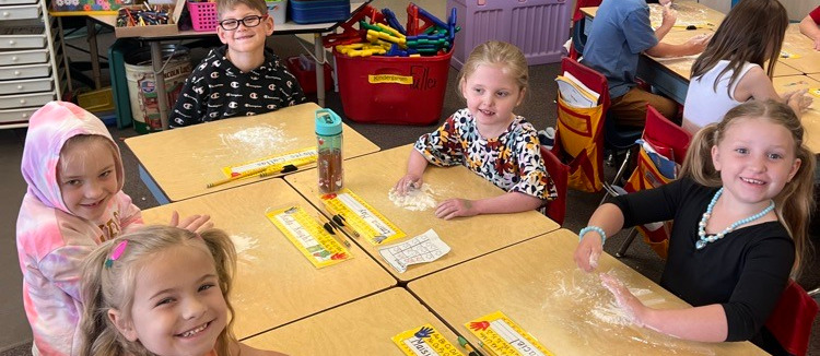Children seated at desks, engaged in an activity. A teacher sits at the back. Bright lighting and wooden cabinets.