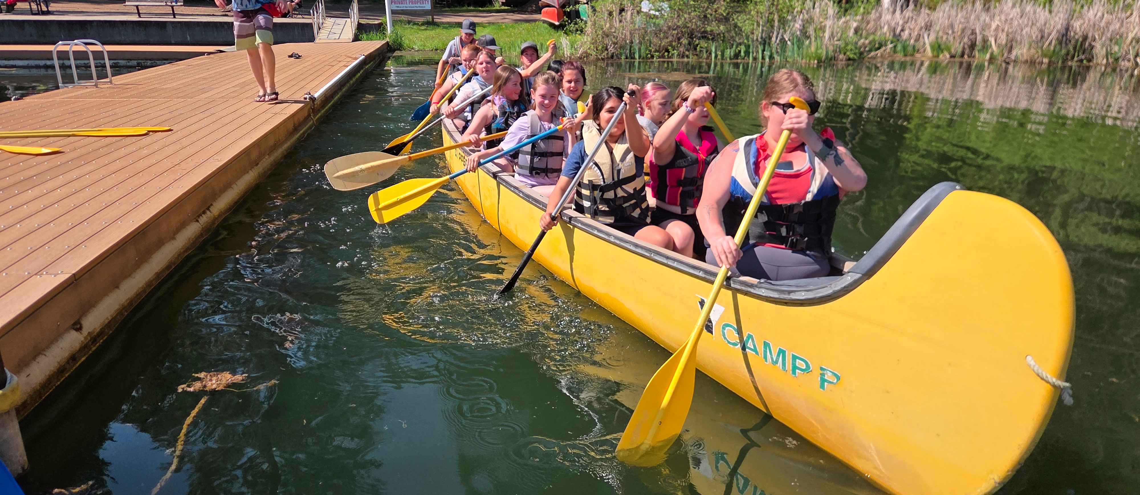 A group of students in life jackets paddling a yellow canoe by a dock with trees in the background.