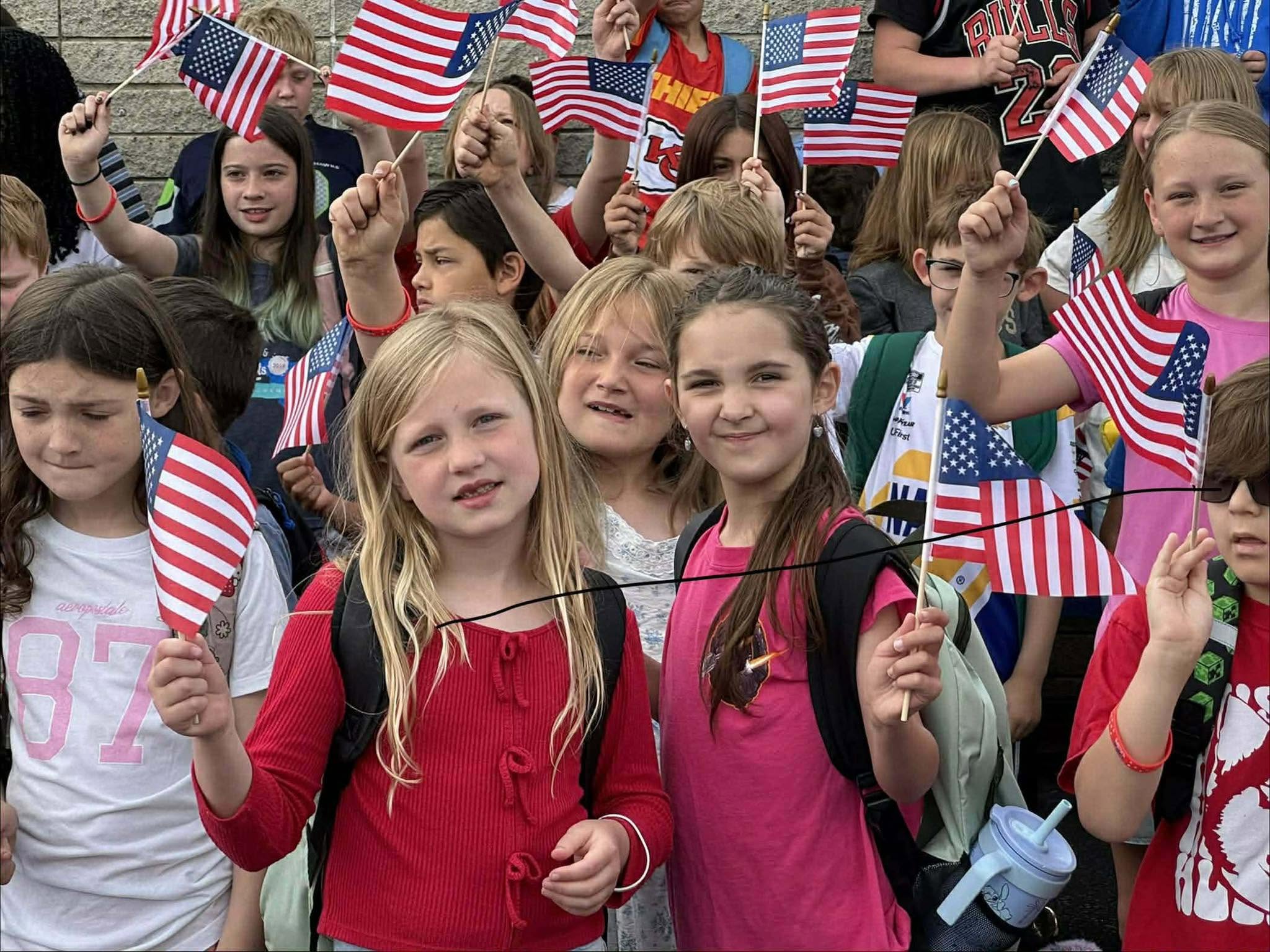 A group of children holding American flags in a school setting, some with backpacks and others carrying flags in different colors.