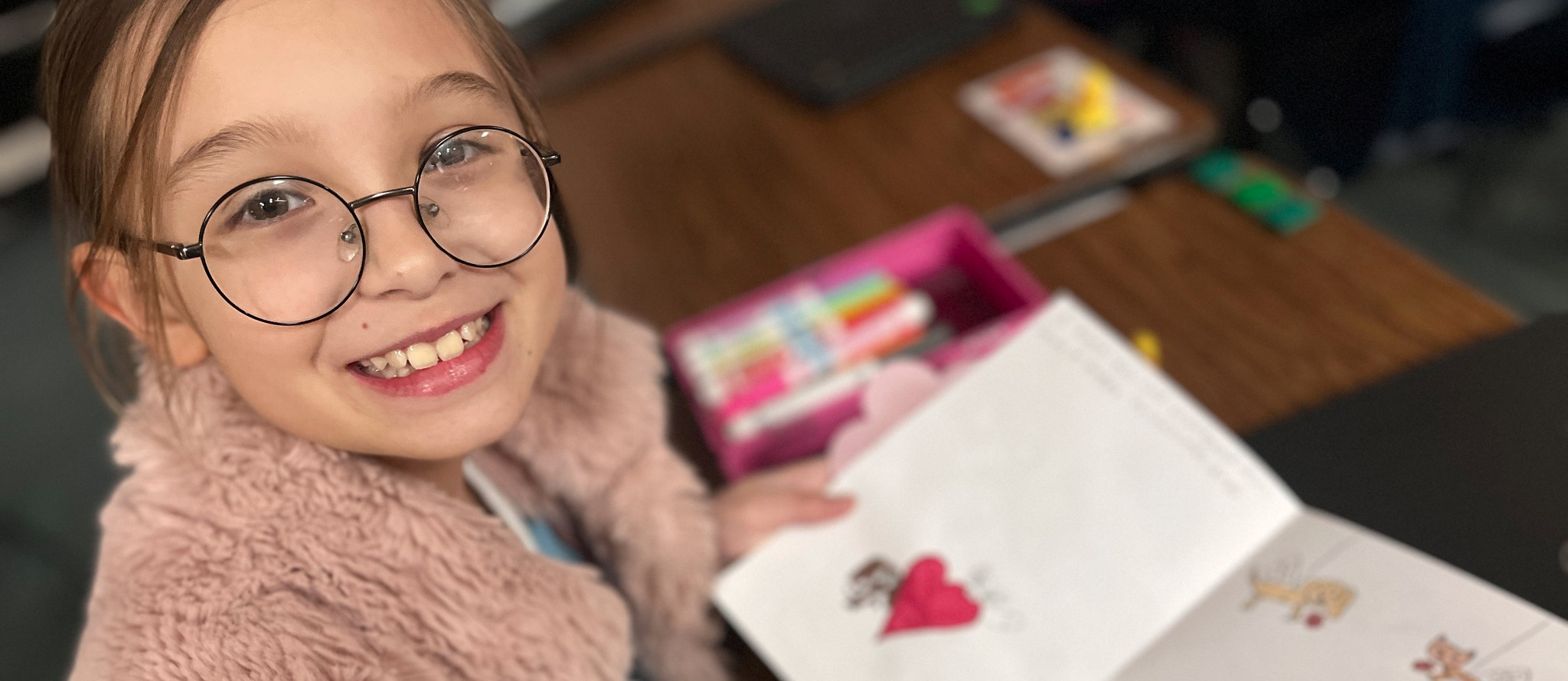 A girl with glasses sits at a desk holding a colorful card with drawings and writing.