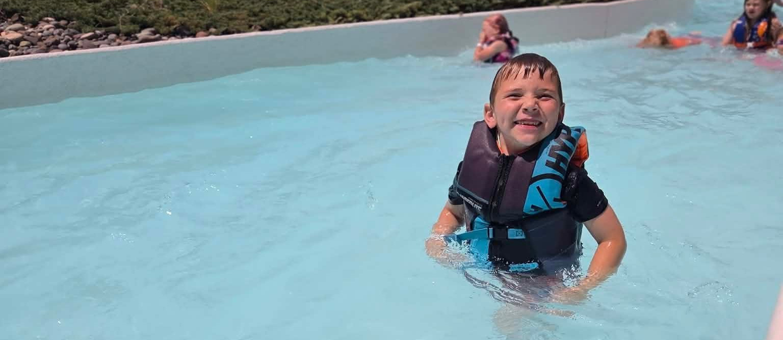 A young person in a blue life jacket in a large pool at a water park.