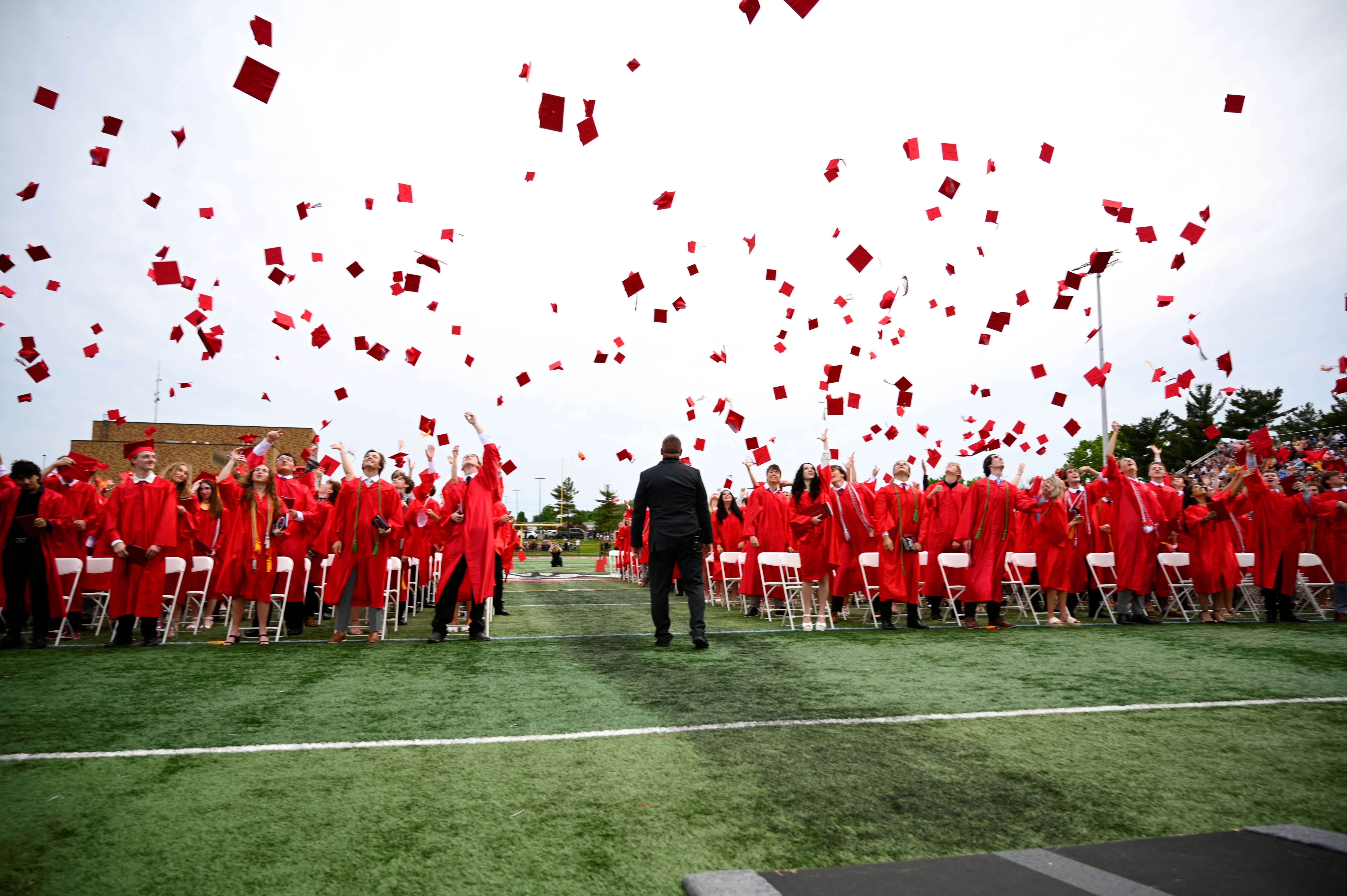 A large group in red gowns throws caps in the air during an outdoor ceremony on a green field.