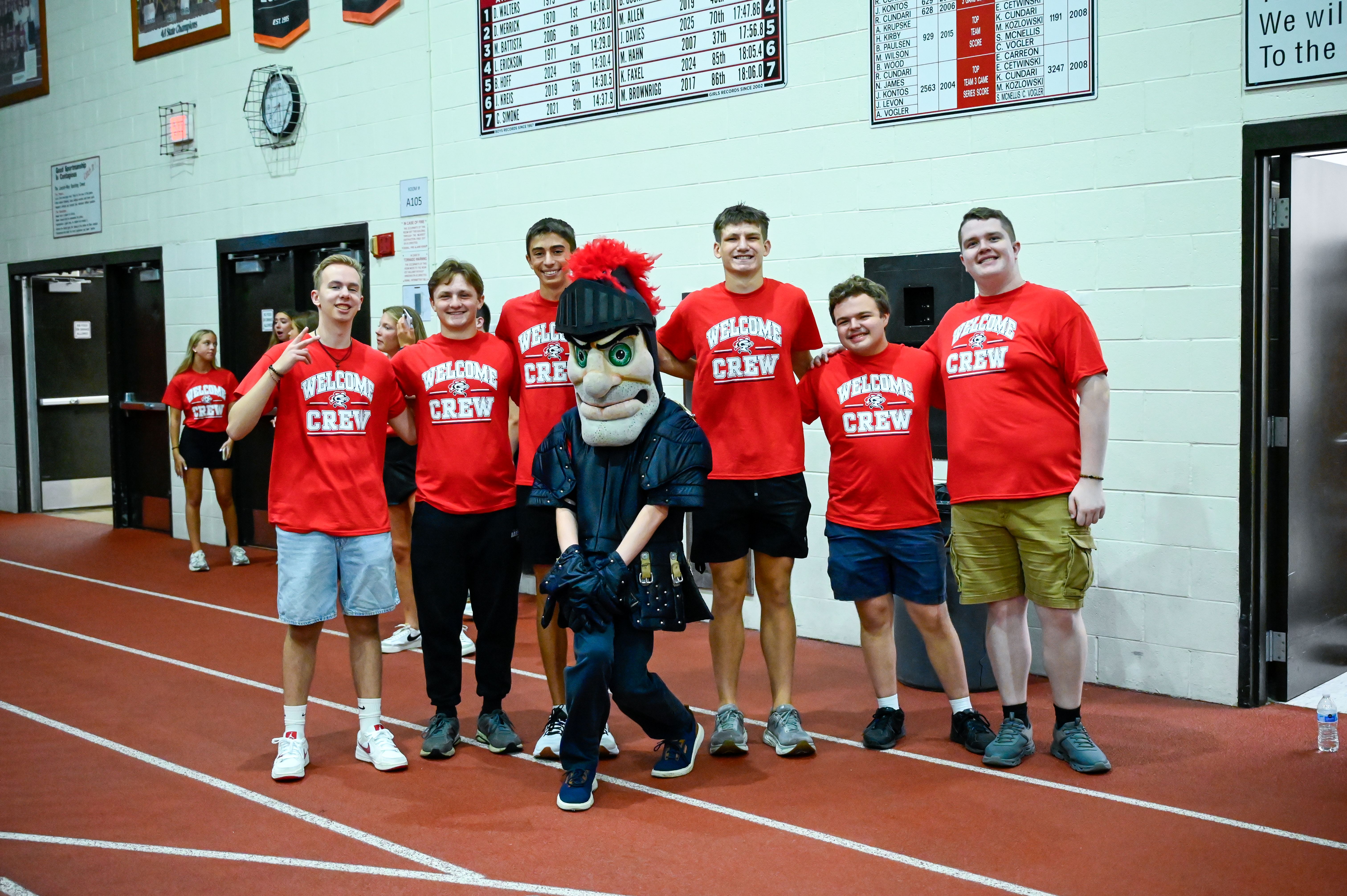 Seven people wearing red shirts pose for a photo on a track, with one person in a mascot costume.