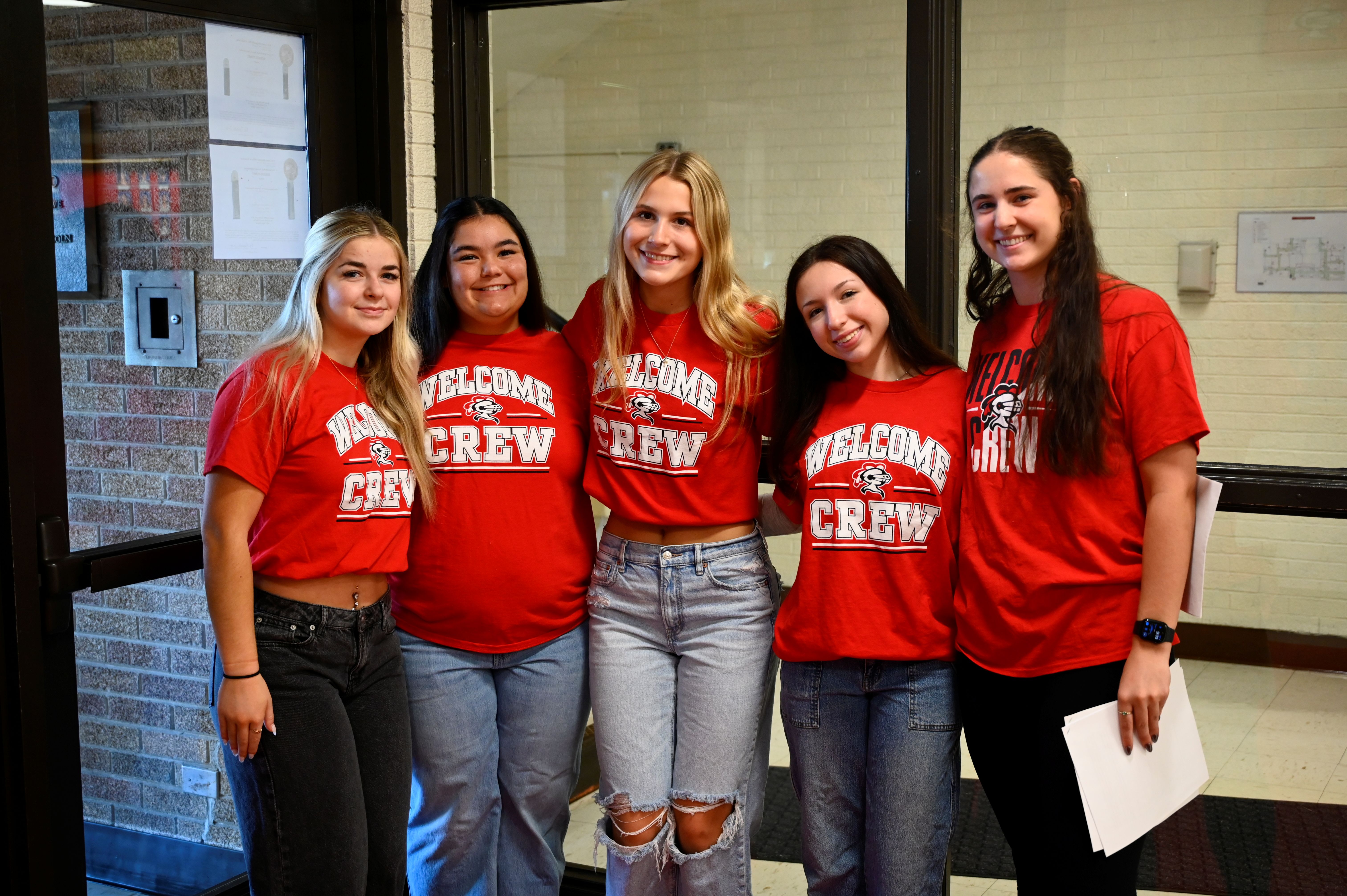 Five girls in red shirts with "Welcome Crew" text pose together in front of a brick wall.
