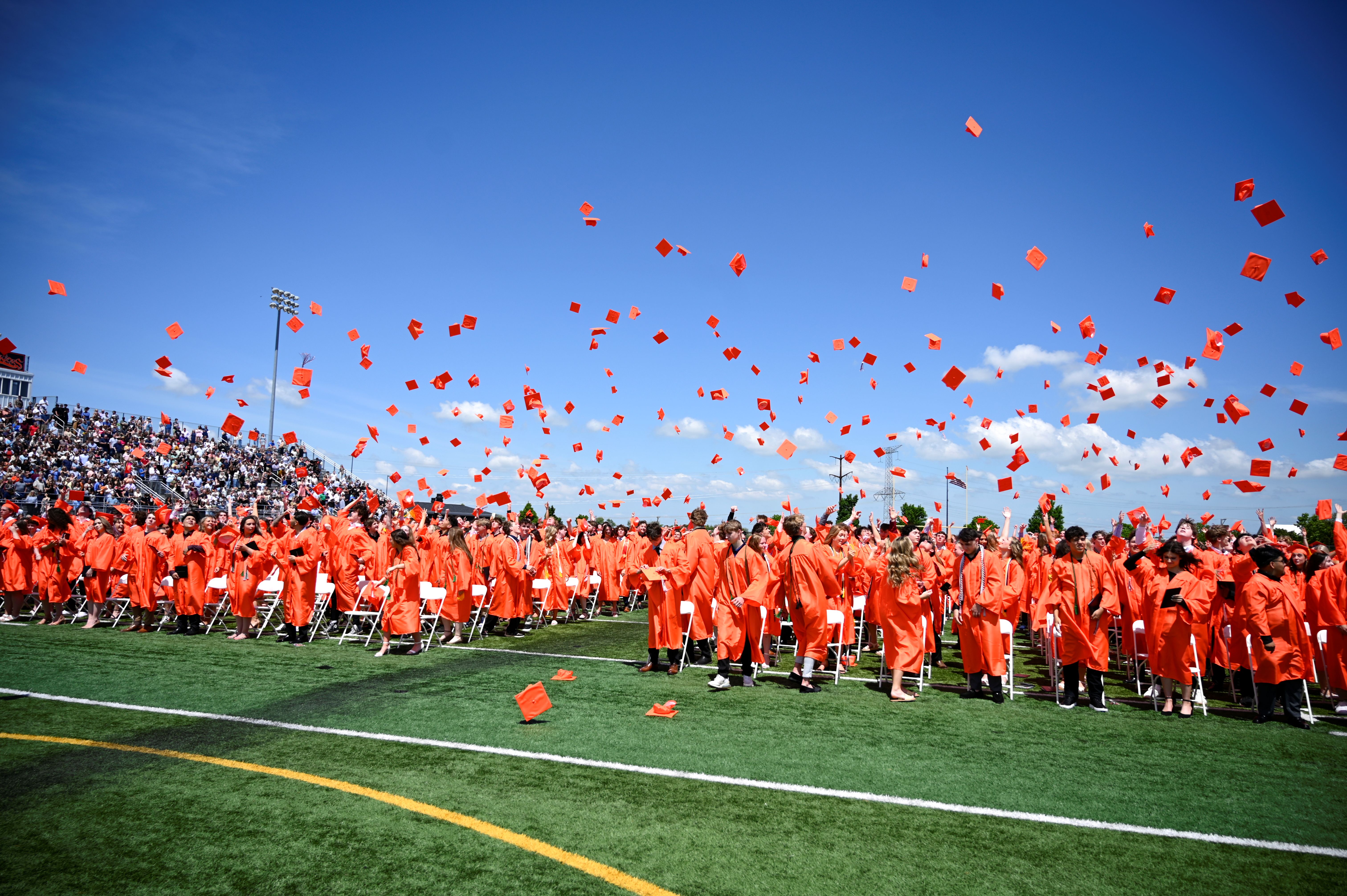 Many people in orange graduation gowns throw red caps into the air on a field with spectators.