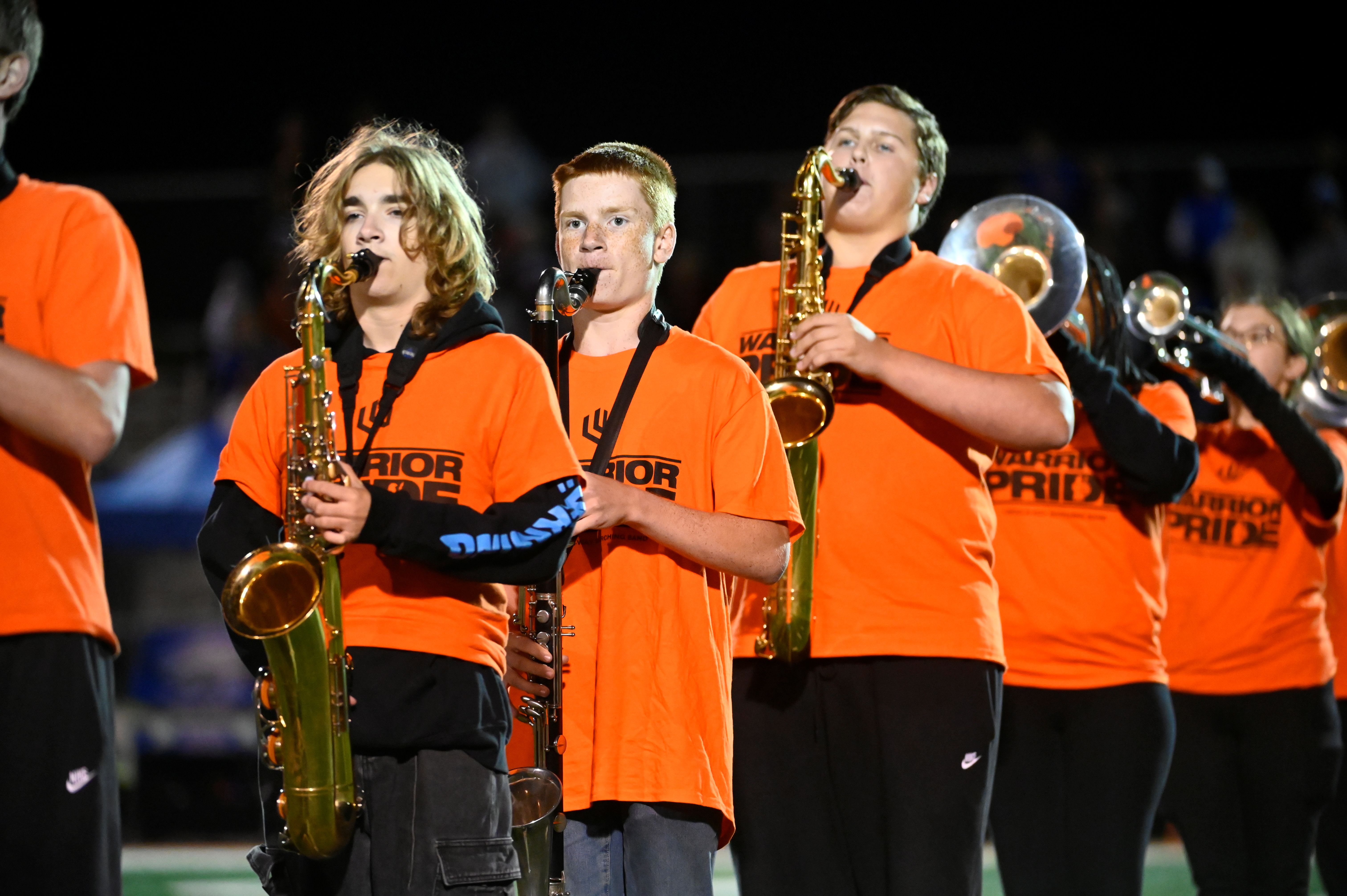 Band students in orange shirts play saxophones and other instruments during an outdoor performance.