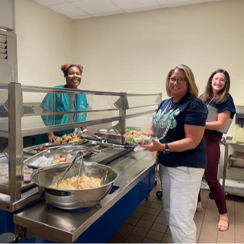 two smiling female teachers trying the new salad bar with a  smiling cafeteria manager behind the bar