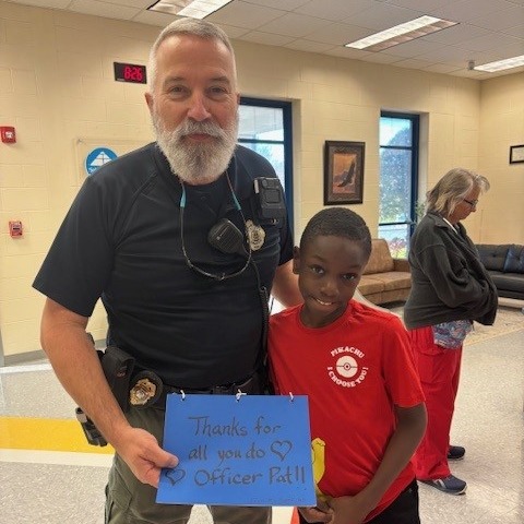 A police officer holding a sign that says "Thanks for all your good deeds." He stands next to a smiling boy.