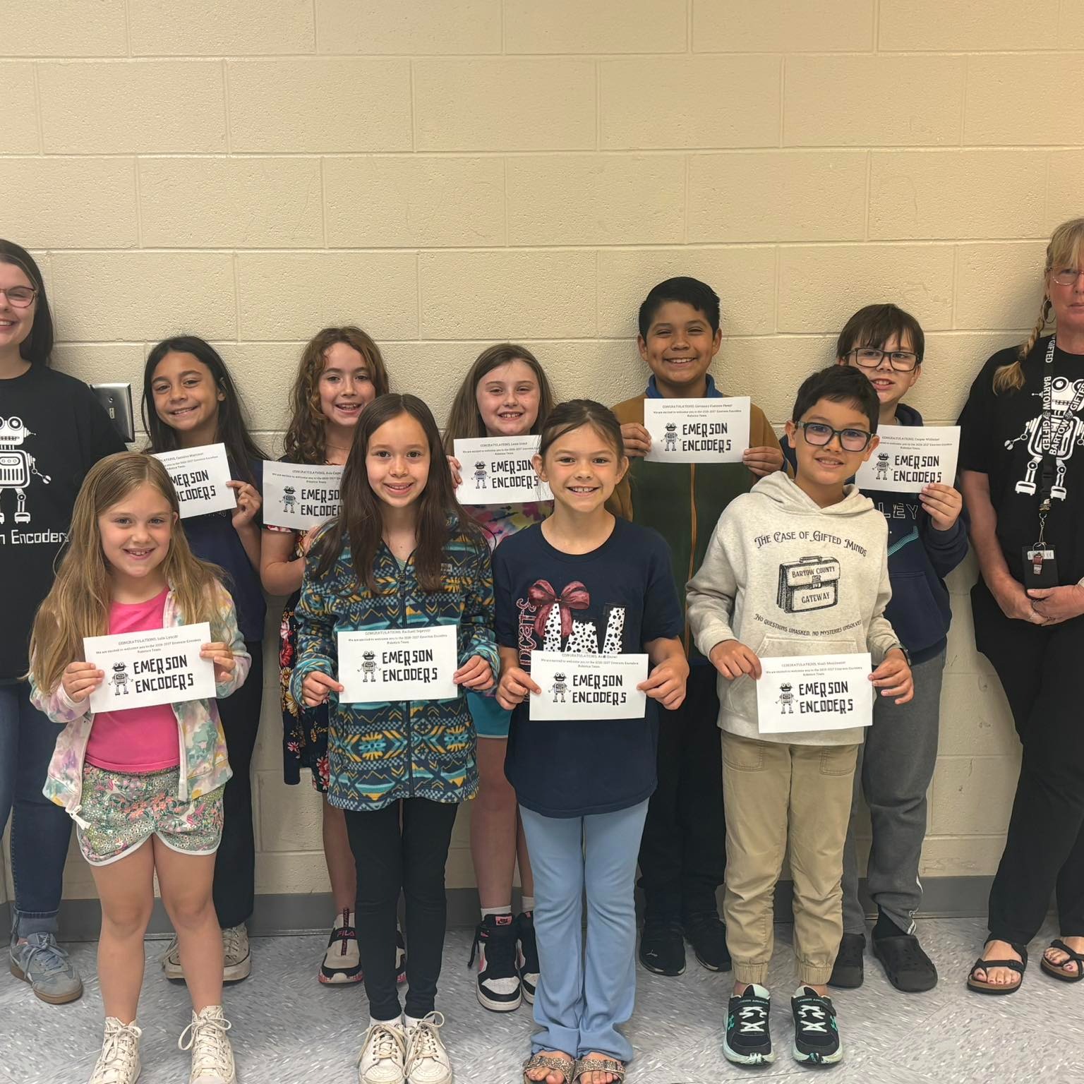 A group of children and adults stand in a row holding signs, with a white wall in the background.