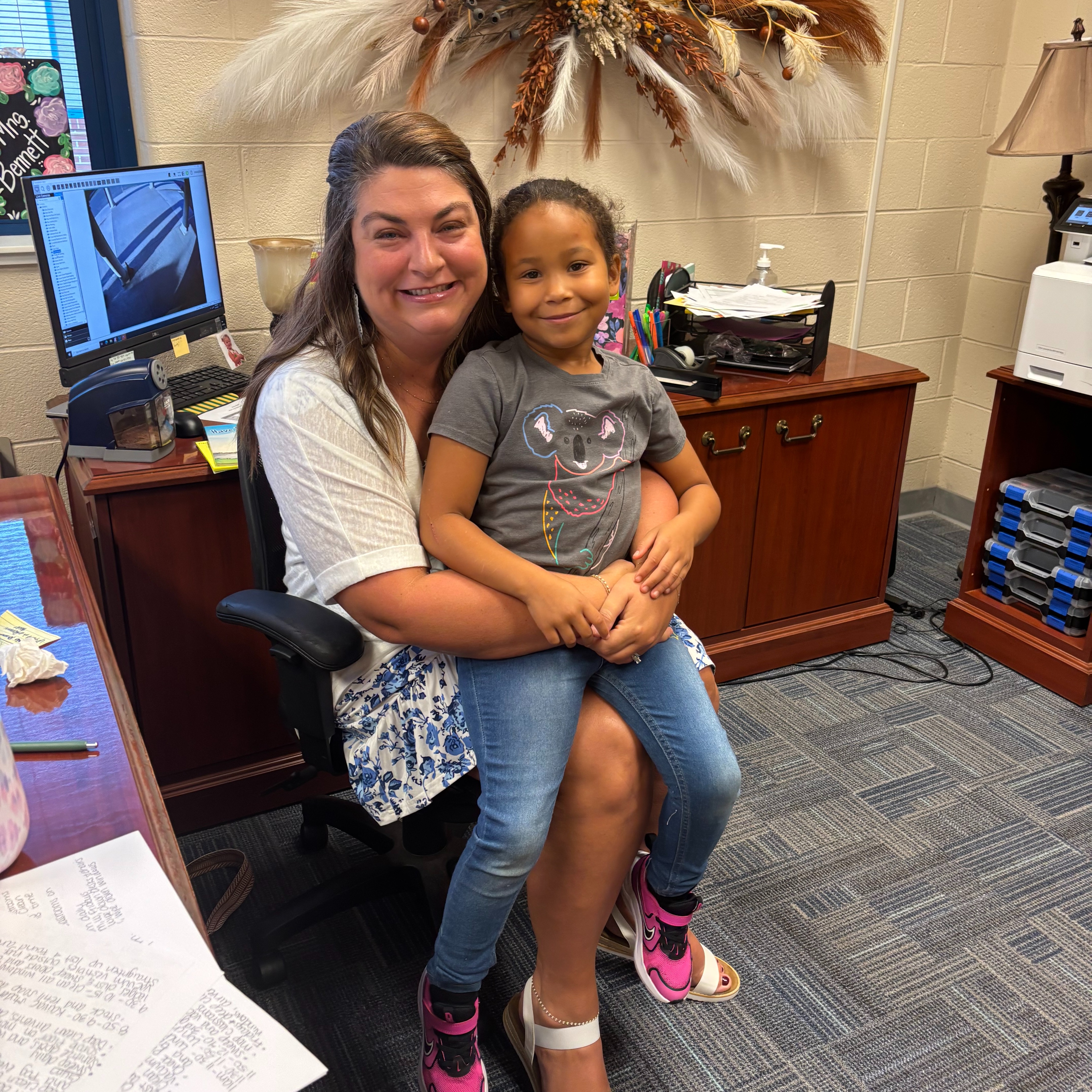 A woman sits in a chair with a child on her lap. They both smile and look at the camera. A desk and lamp are in the background.