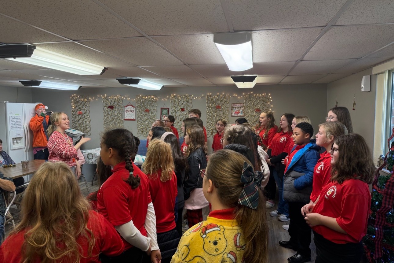 A group of students in red shirts and ponytails stand in a room with a speaker.