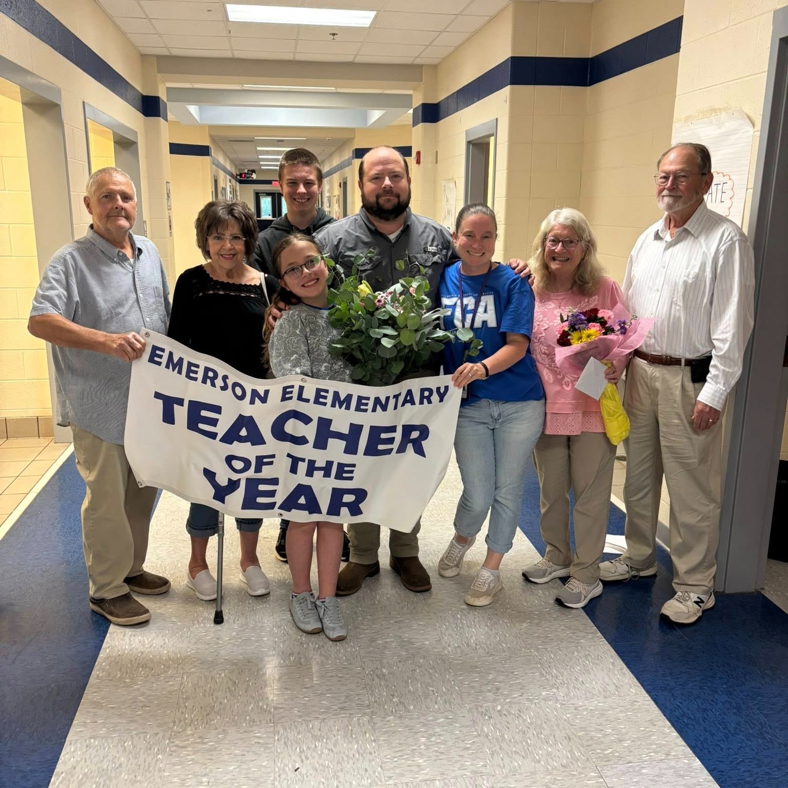A group of people stand in a hallway, one person holding a sign that reads "Teacher of the Year."