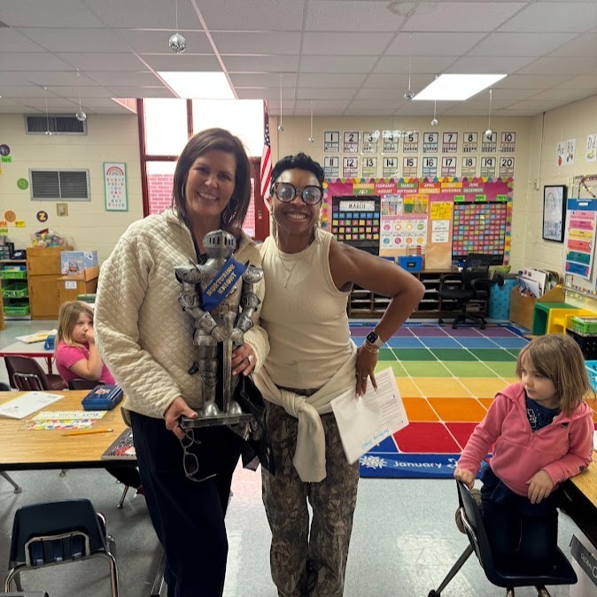 Two adults and a child are in a classroom. One adult holds a trophy, another holds paper. Colorful rug on floor.