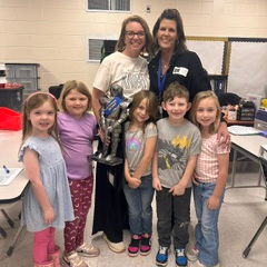 Group of children and two women stand together in a classroom, smiling. One child holds a trophy.