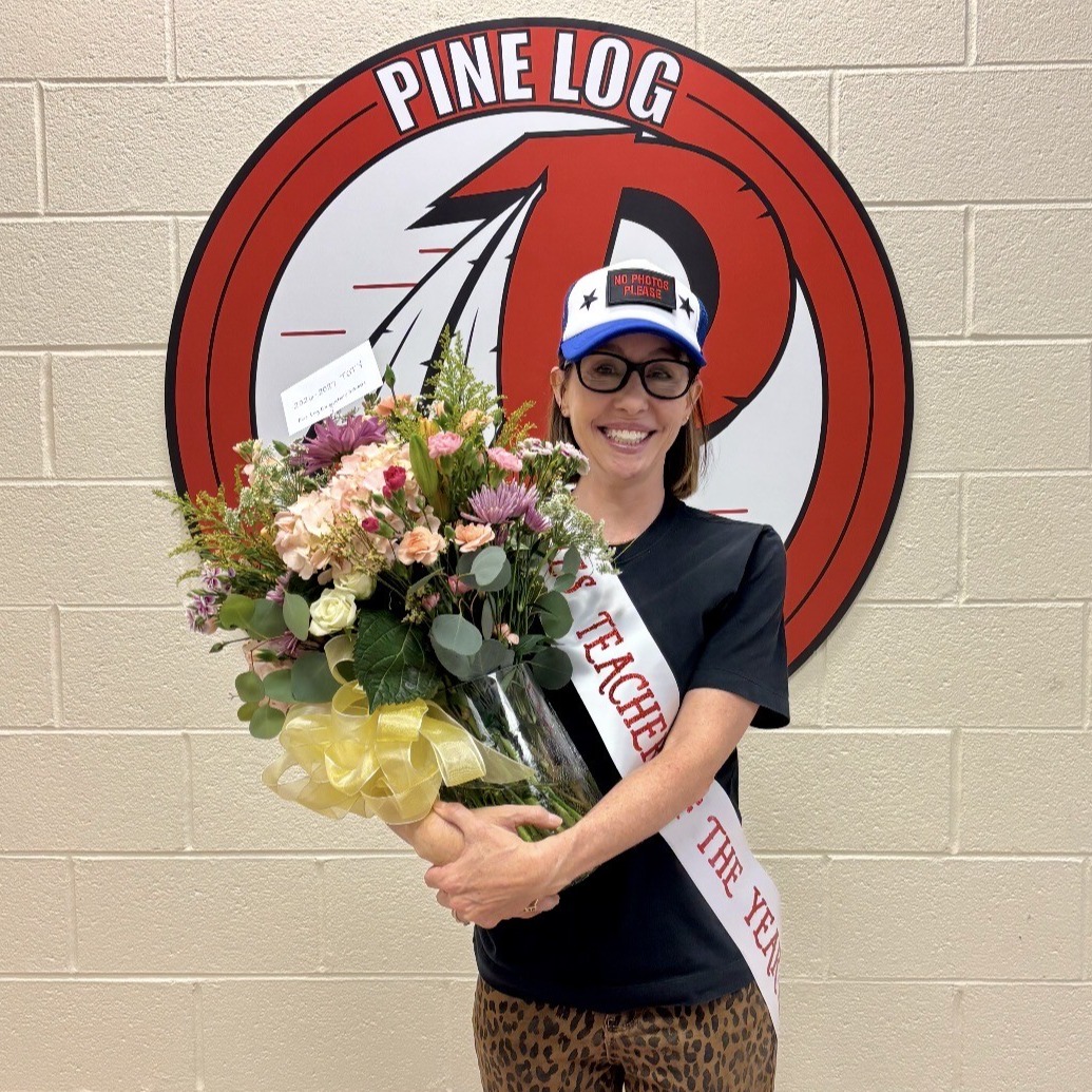 A woman stands holding a bouquet of flowers. She wears a black t-shirt and leopard-print pants. Behind her is a wall with a logo.