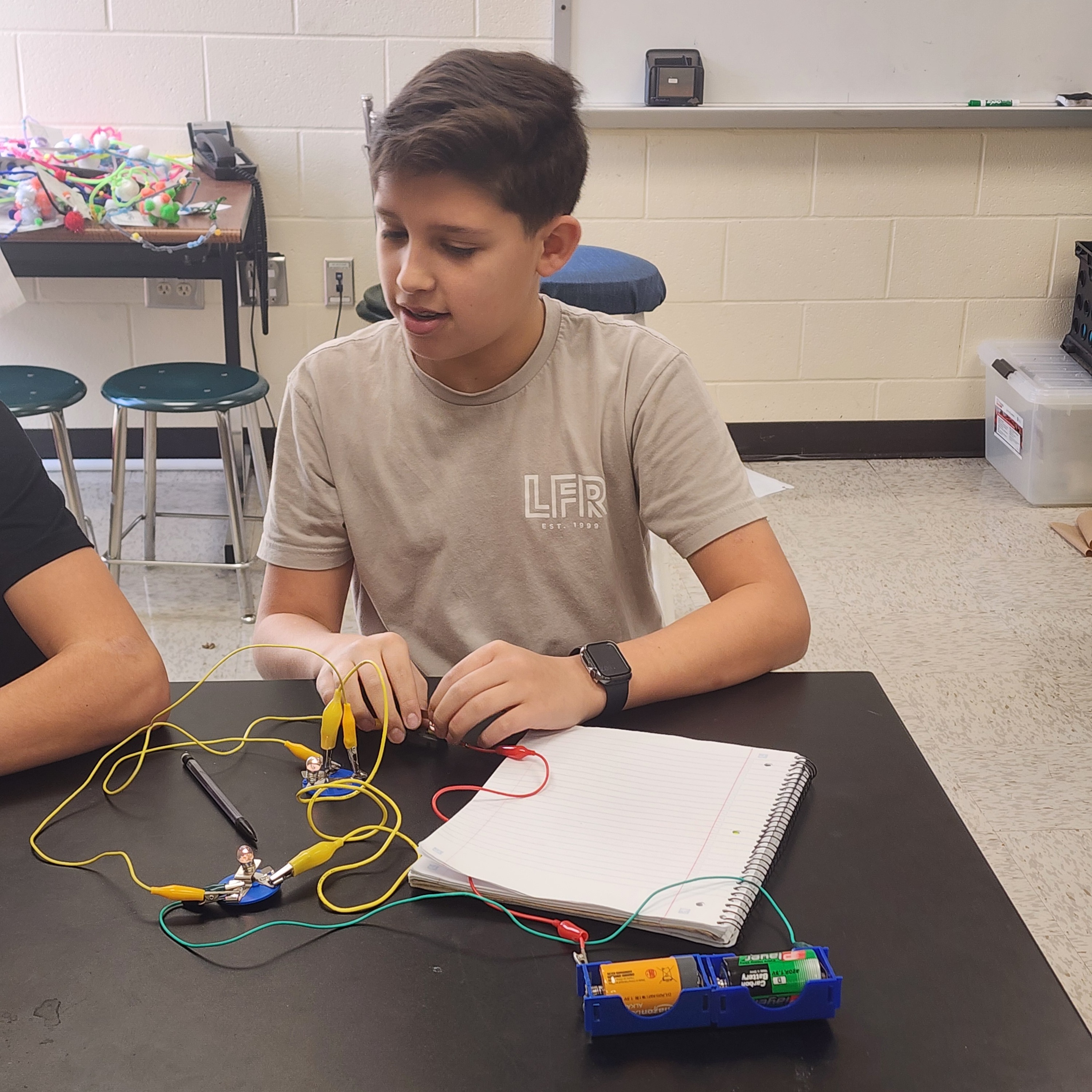 A boy is sitting at a desk, working with a laptop, wires, and a circuit board.