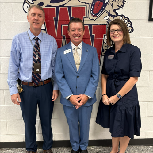 Three individuals stand in front of a wall featuring a school mascot. Two men in suits and one woman in a dress.