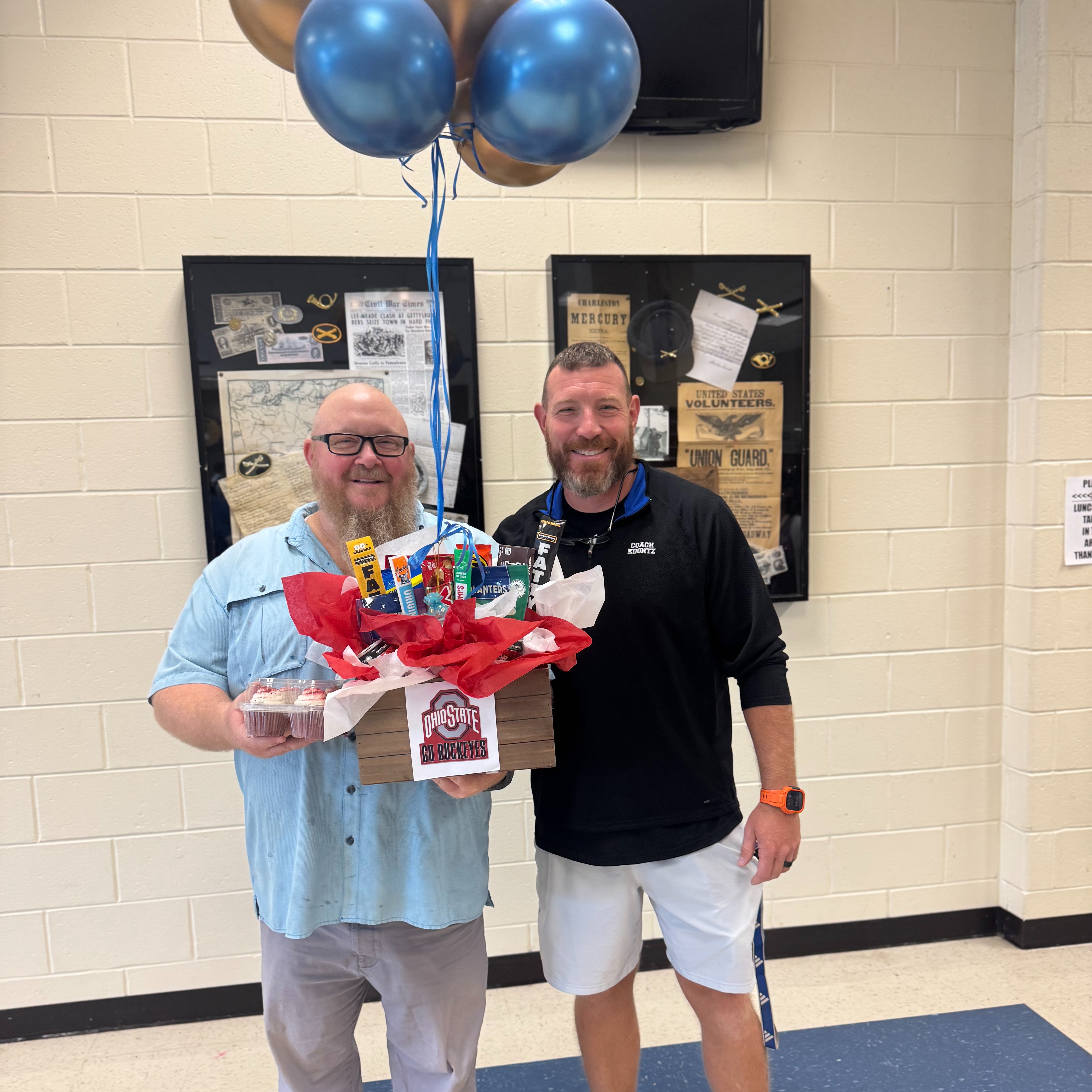 Two men stand next to each other, one holding a basket with snacks and balloons, and the other smiling. They are indoors with a white wall and bulletin board behind them.
