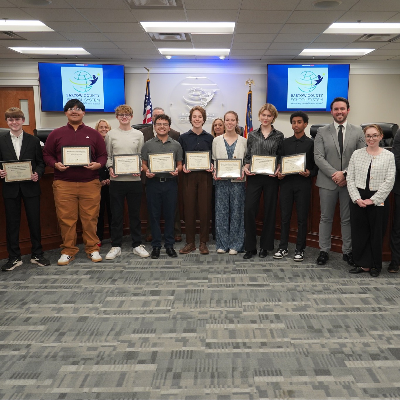 Group of young people with certificates stand in a room with flags, a podium, and monitors.