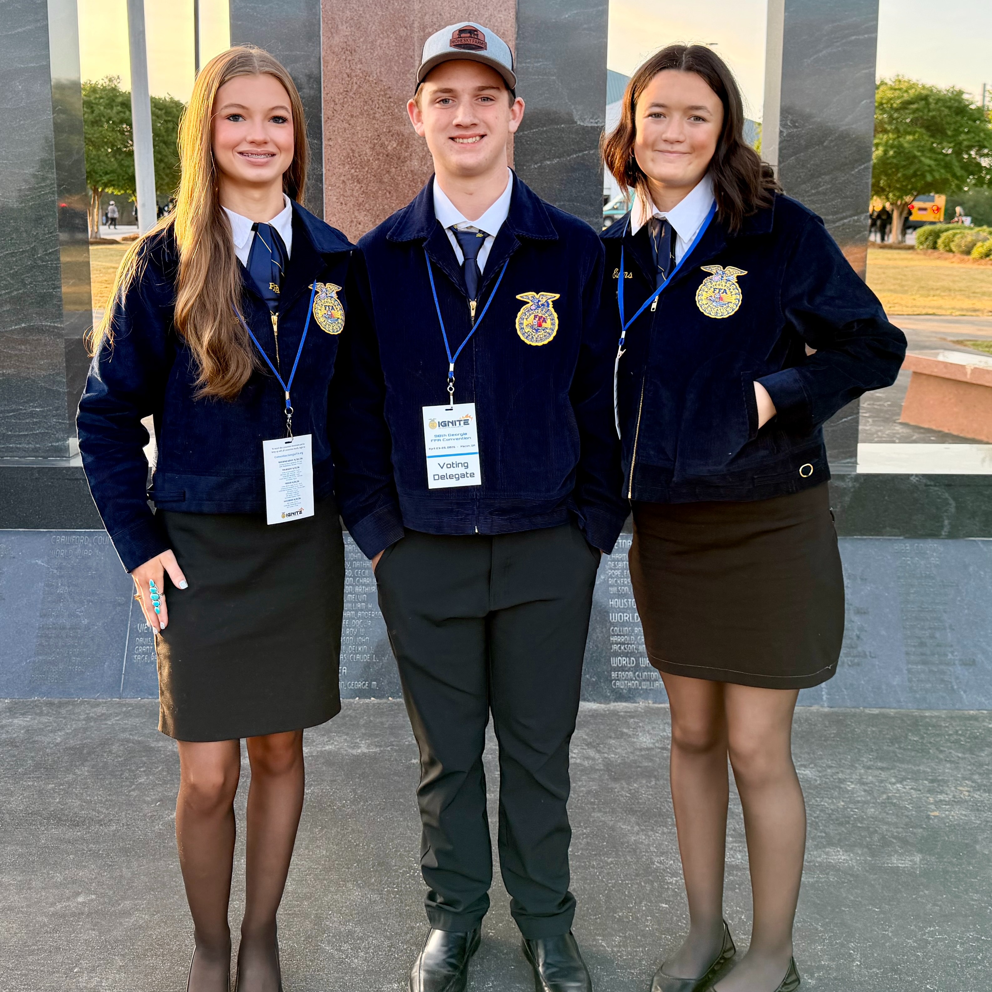 Three students wearing matching jackets and ID lanyards stand together, smiling for a photo in front of a stone monument.