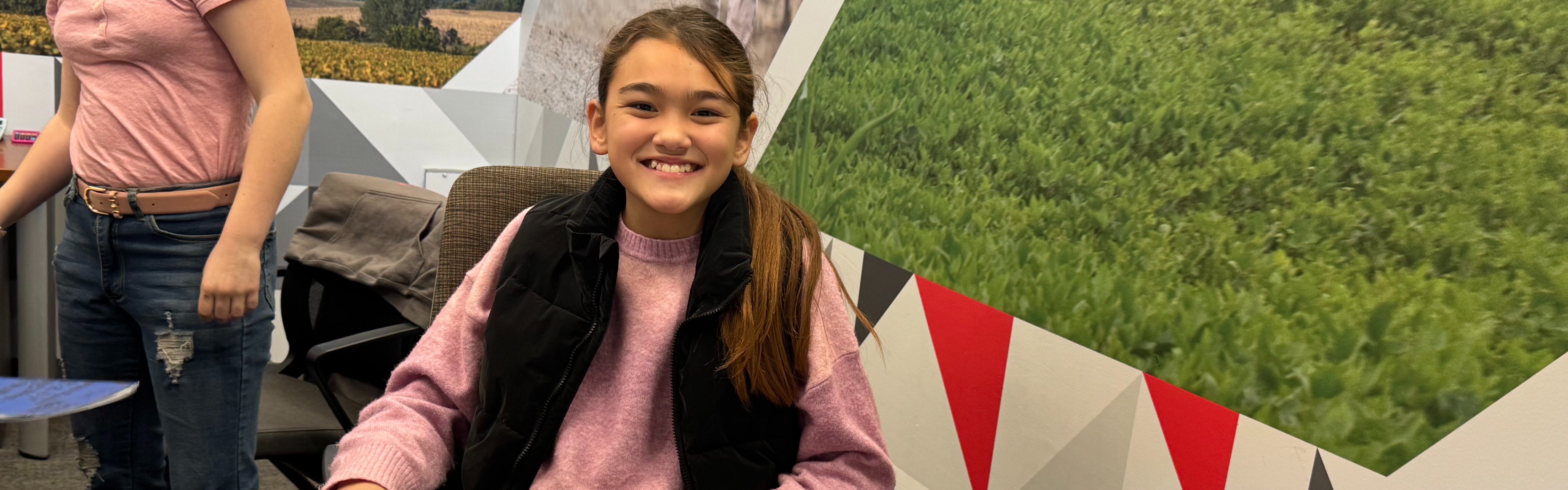 Image of a female 6th grade student sitting at a business desk in front of a laptop smiling at the camera