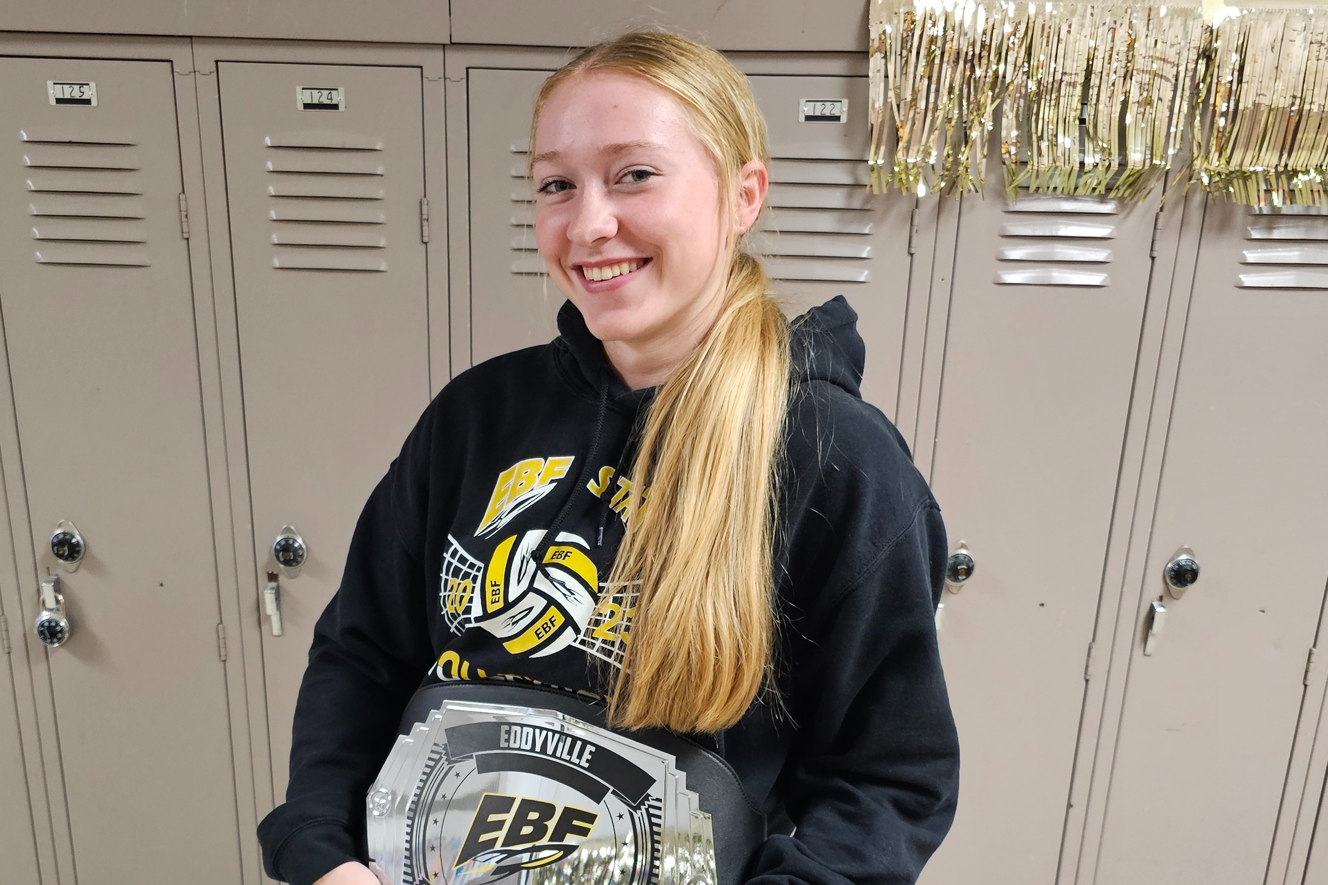 Student poses with the EBF Student of the Week belt in a school hallway lined with lockers. The student is wearing a black EBF volleyball sweatshirt, and gold streamers decorate the area above the lockers.