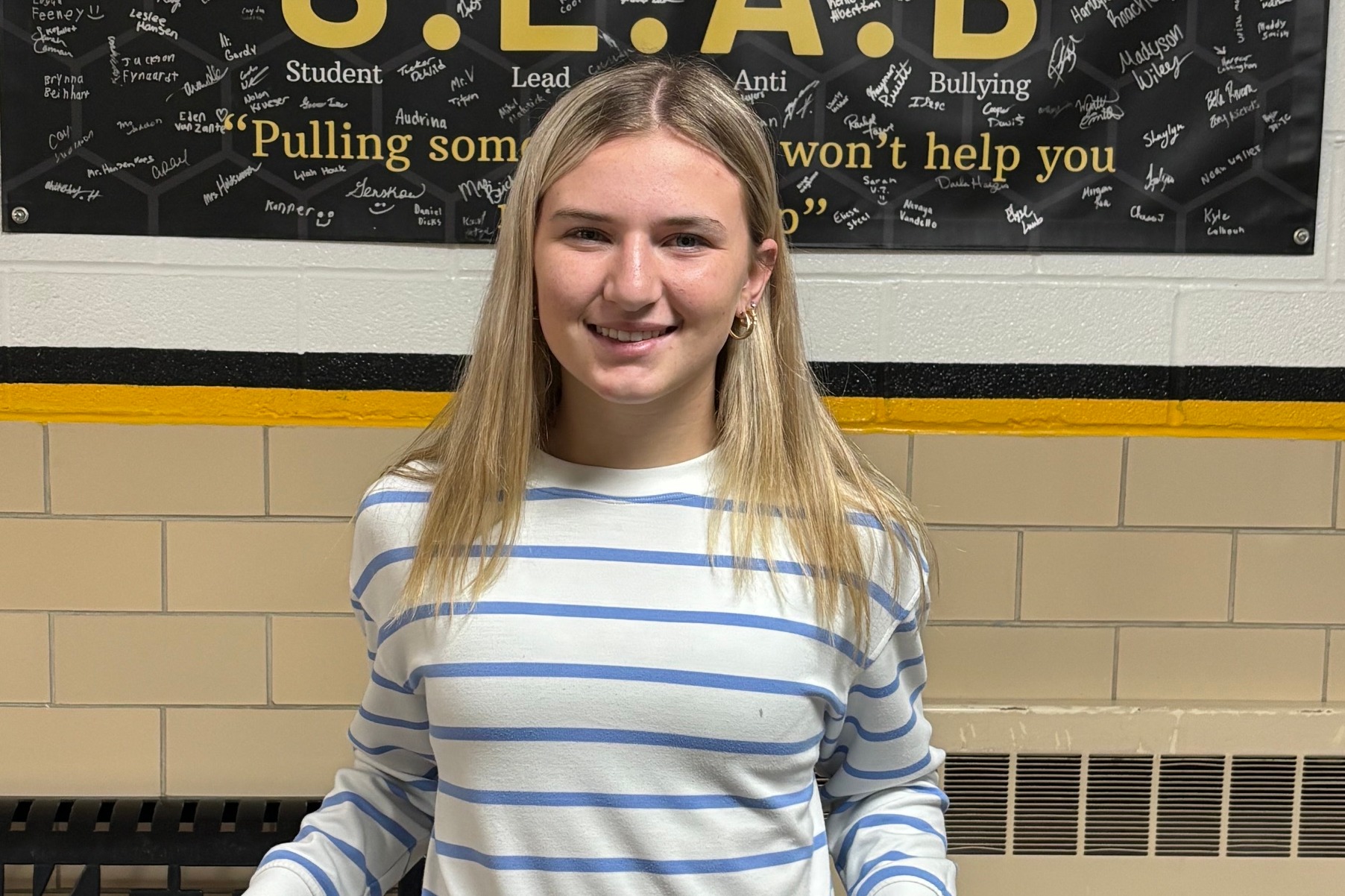 Jr./Sr. High Student of the Week posed for a portrait against a school hallway wall with black and gold accent stripes.