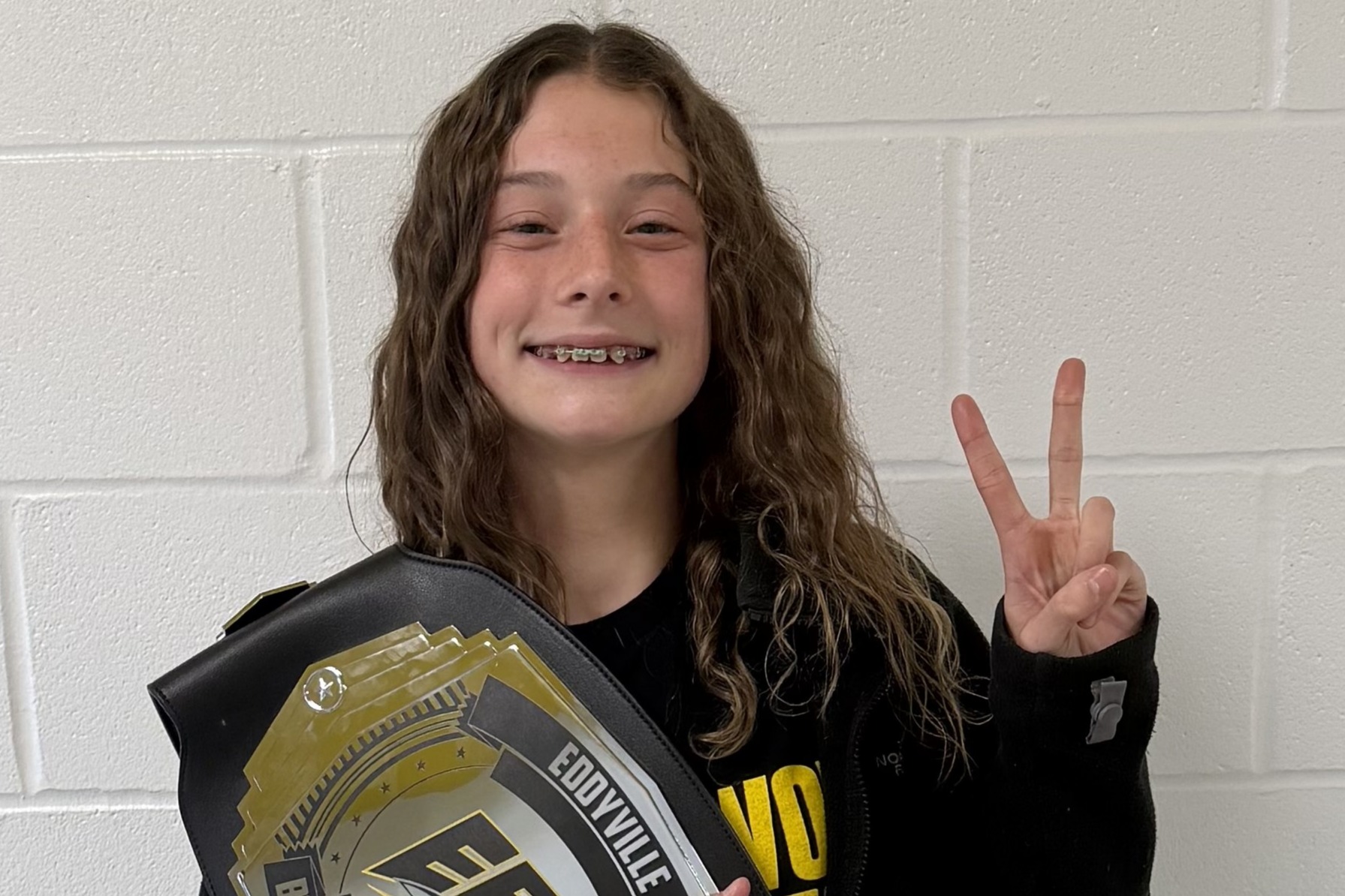 Student poses with the EBF Student of the Week belt in a school hallway lined with lockers. The student is wearing a black EBF volleyball sweatshirt, and gold streamers decorate the area above the lockers.