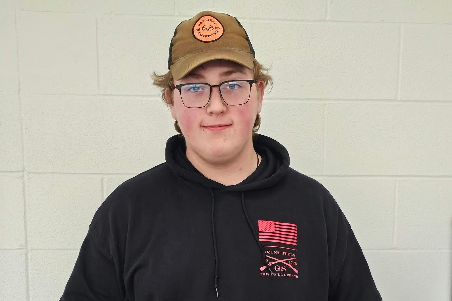 A student holds a large belt trophy against a white wall, wearing a hat, glasses, and a black hoodie.