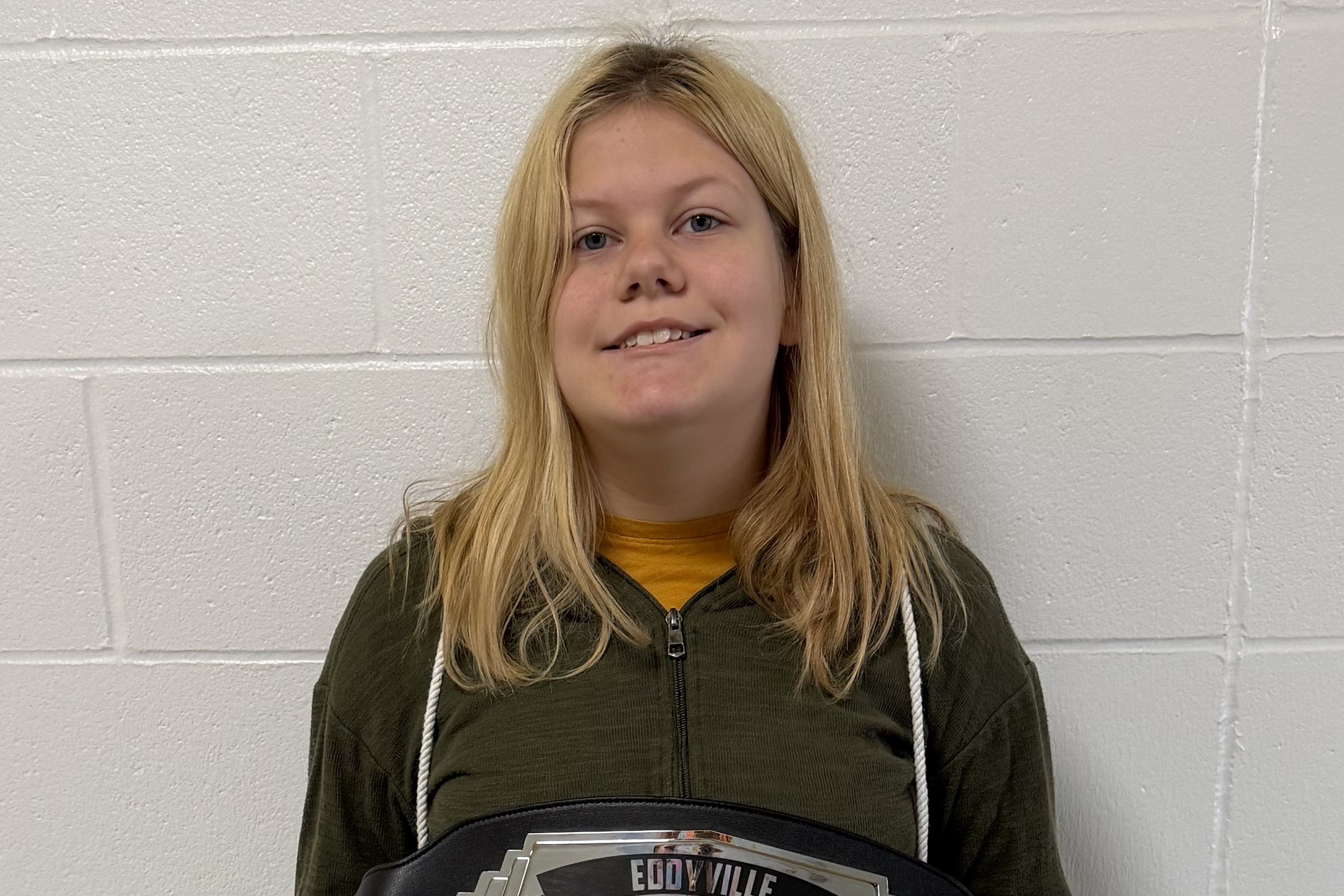 A girl holds a championship belt around her waist, smiling. She wears a green hoodie.