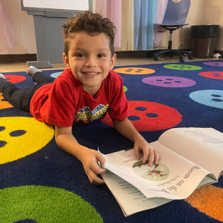 Preschool boy with red shirt on laying on a carpet reading a book