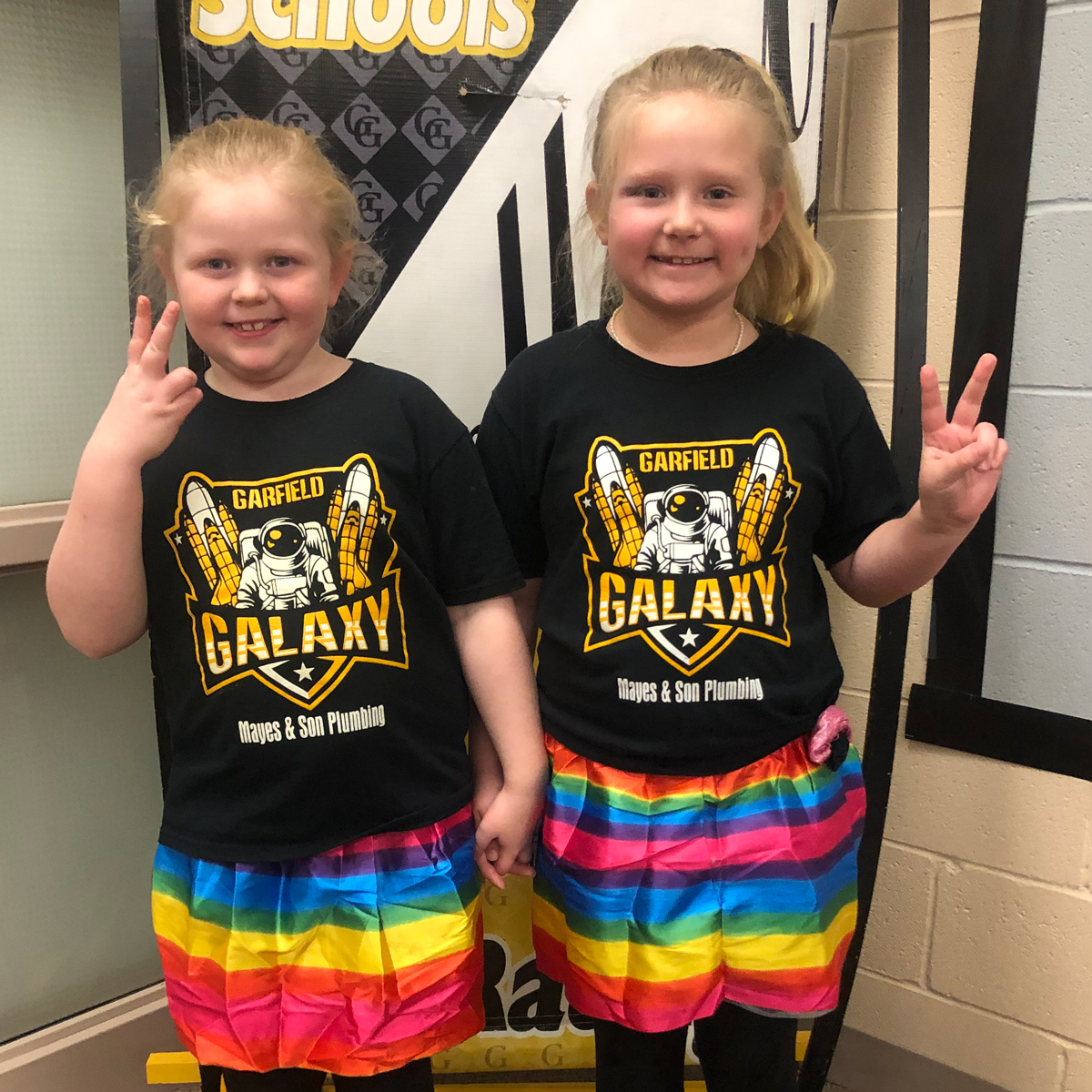 Two elementary students wearing black and gold shirts stand in front of the Excellence banner.