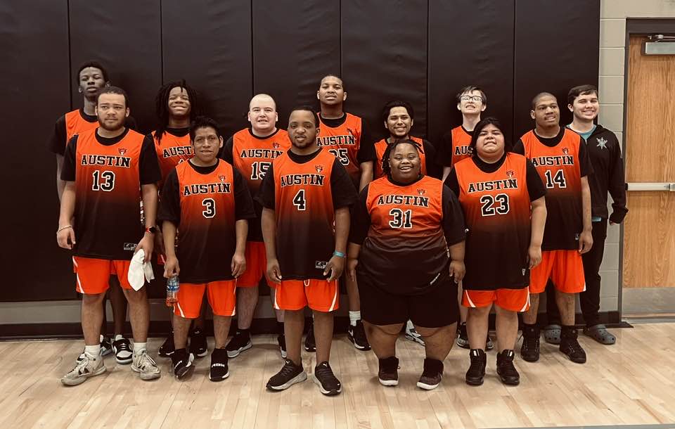 A medium, eye-level team photo of eleven basketball players from the Austin AIM program standing together on an indoor gym court. They are wearing matching black and orange gradient athletic jerseys with orange shorts, featuring 'AUSTIN' printed on the front above their respective jersey numbers. The players are posing with smiles against a background of solid black gym wall padding, with a male coach or support staff member smiling on the far right.