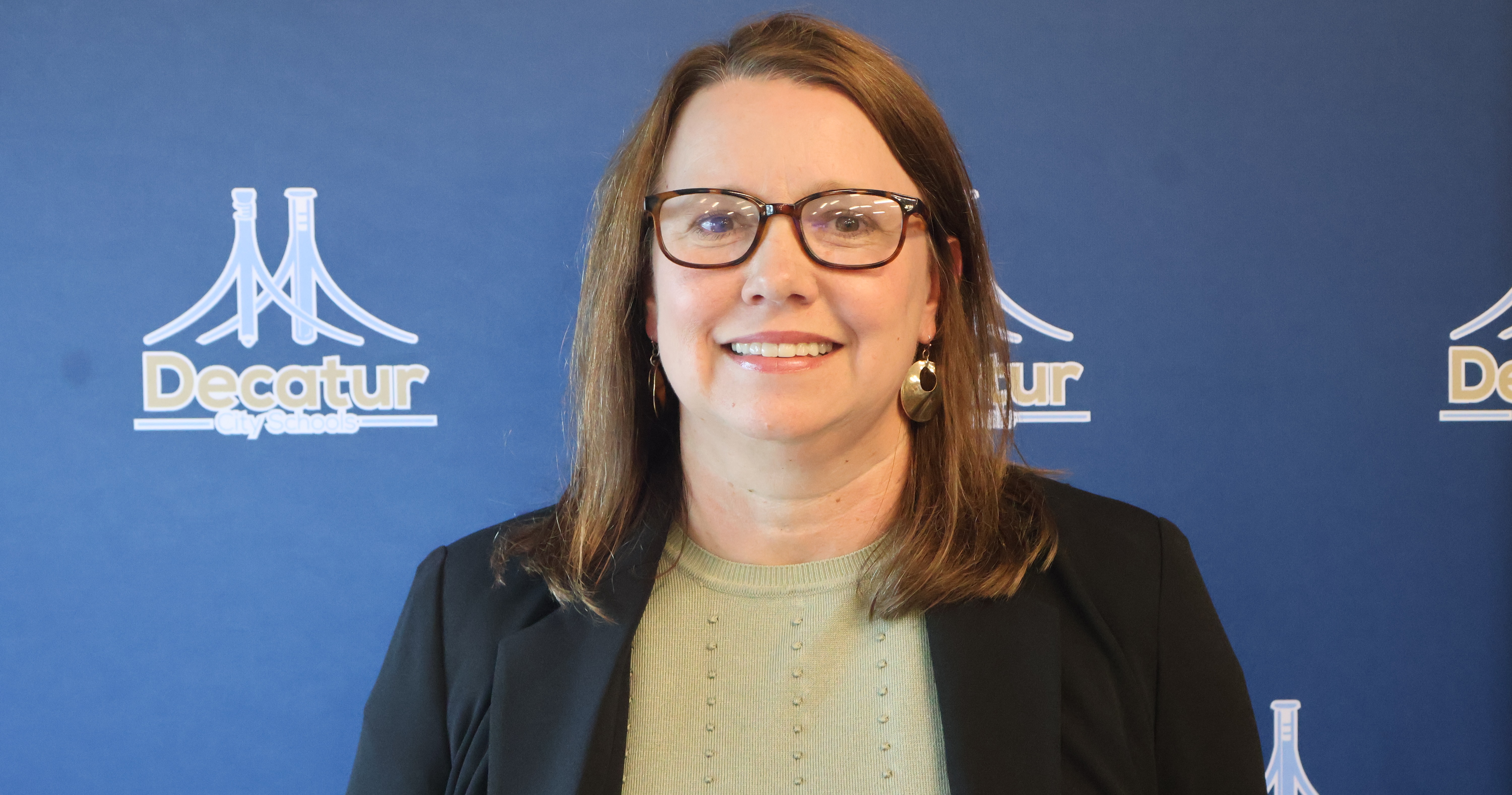 A professional, medium headshot of Mrs. Wendy Henderson, the new principal at Decatur Middle School. She is a woman with shoulder-length brown hair, wearing tortoiseshell glasses, a light sage green top, dangling gold earrings, and a black blazer jacket. She is smiling warmly at the camera against a blue backdrop featuring the white Decatur City Schools logo.