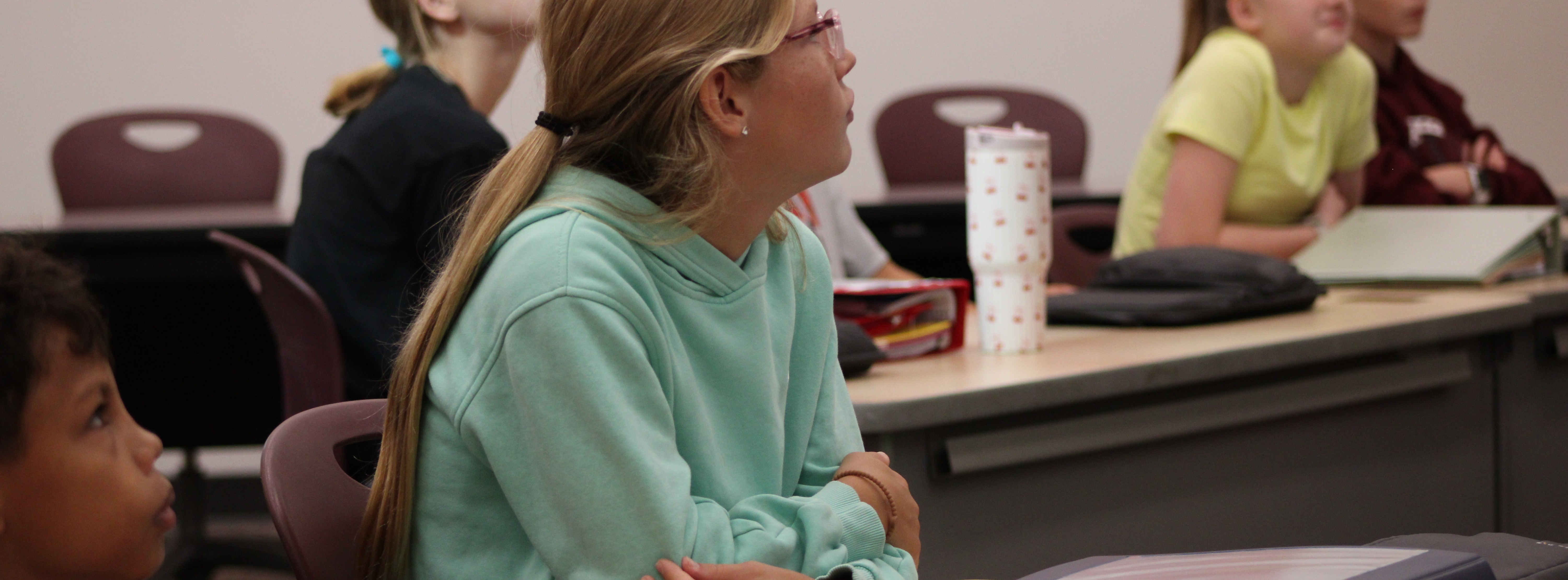 A group of students sit in a classroom. A girl in green looks upwards. Another student sits nearby.