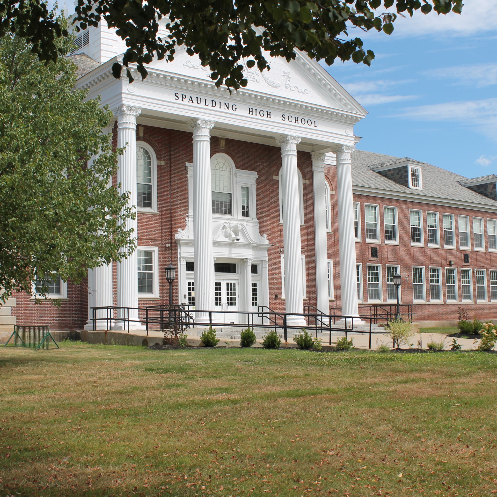 A large school building with white columns and a gray roof, surrounded by a grassy area and trees.