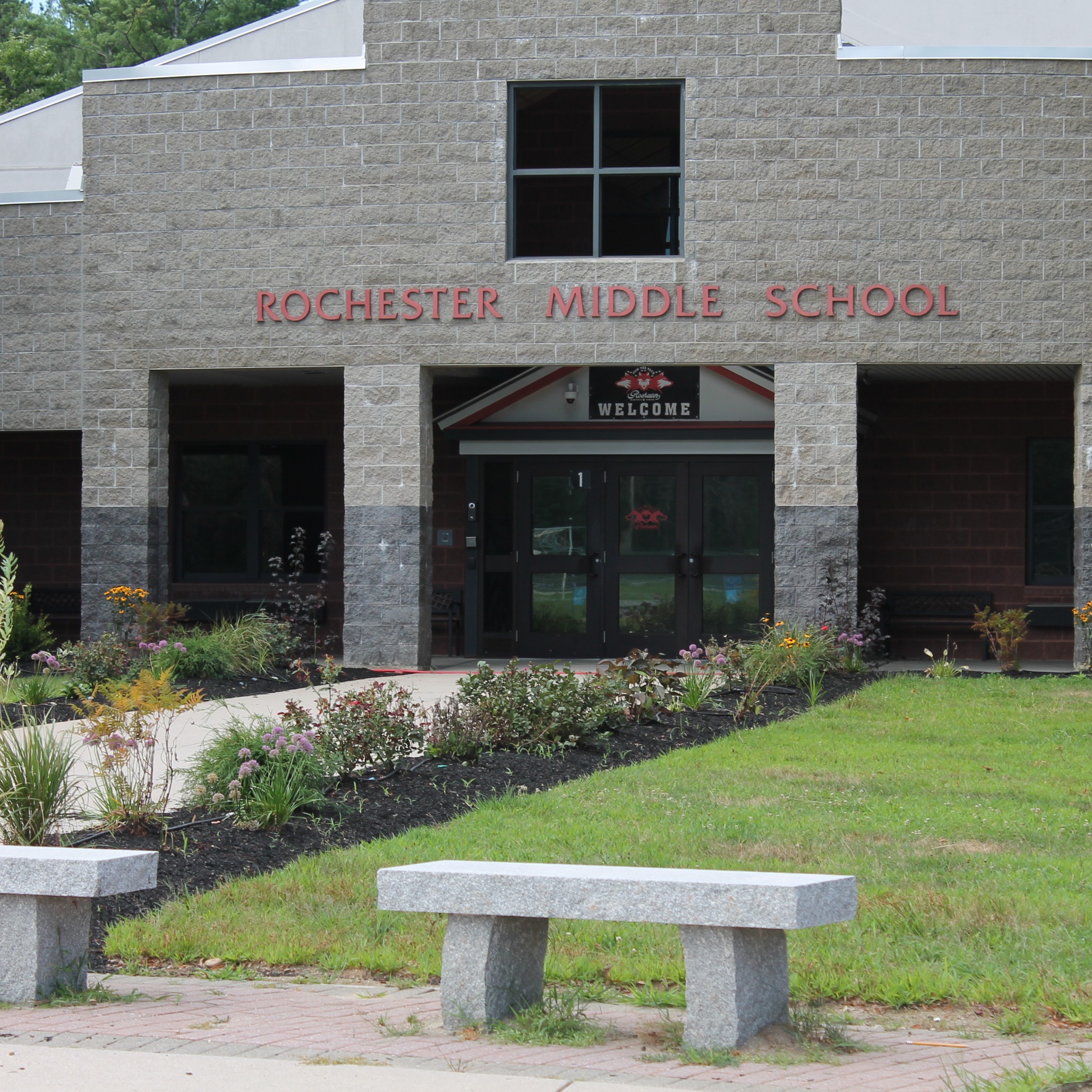 Rochester Middle School building with red lettering. Two benches sit on the lawn in front of the building.