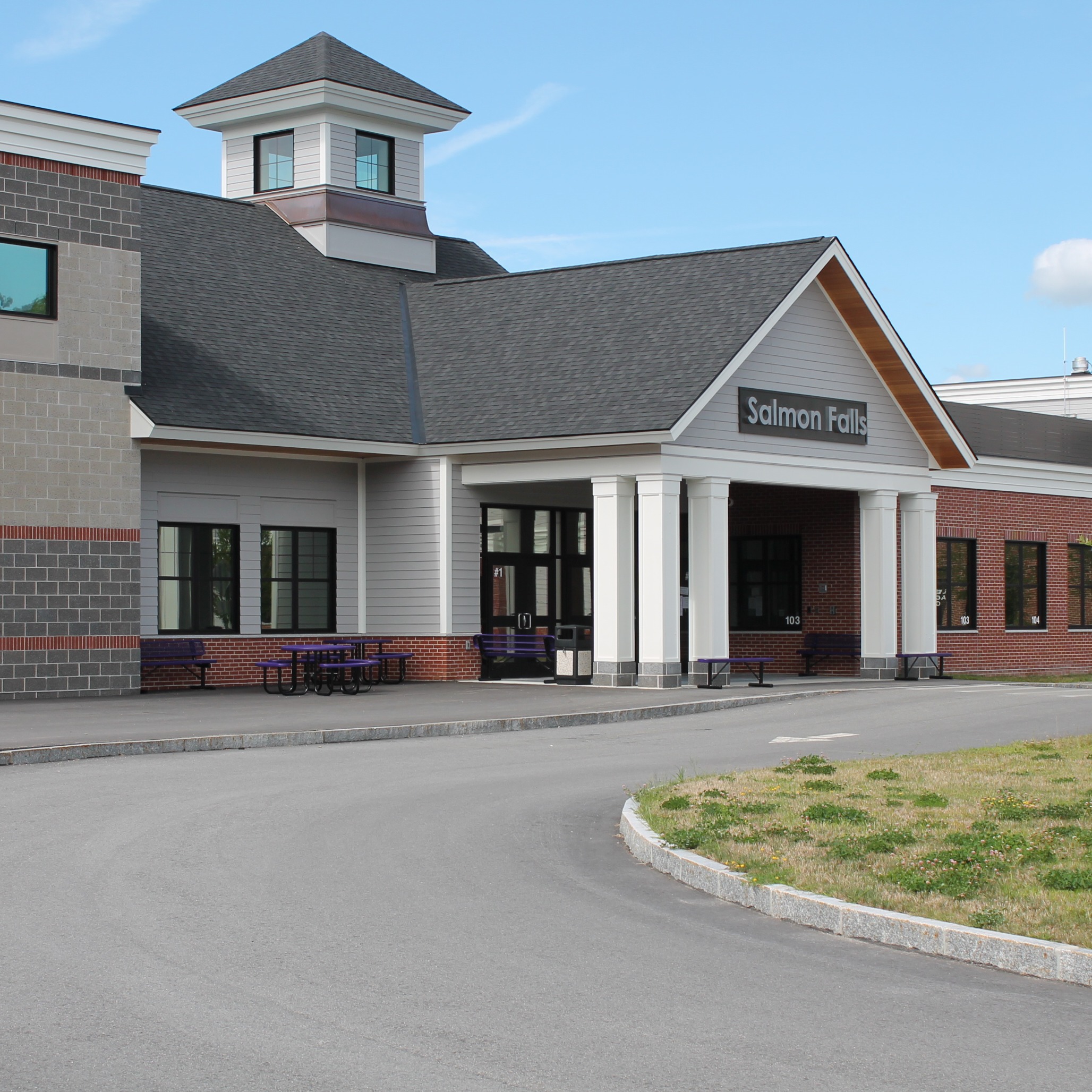 Front entrance of a school building with a gray roof and white pillars. Windows line the walls, and a grassy area is to the right.