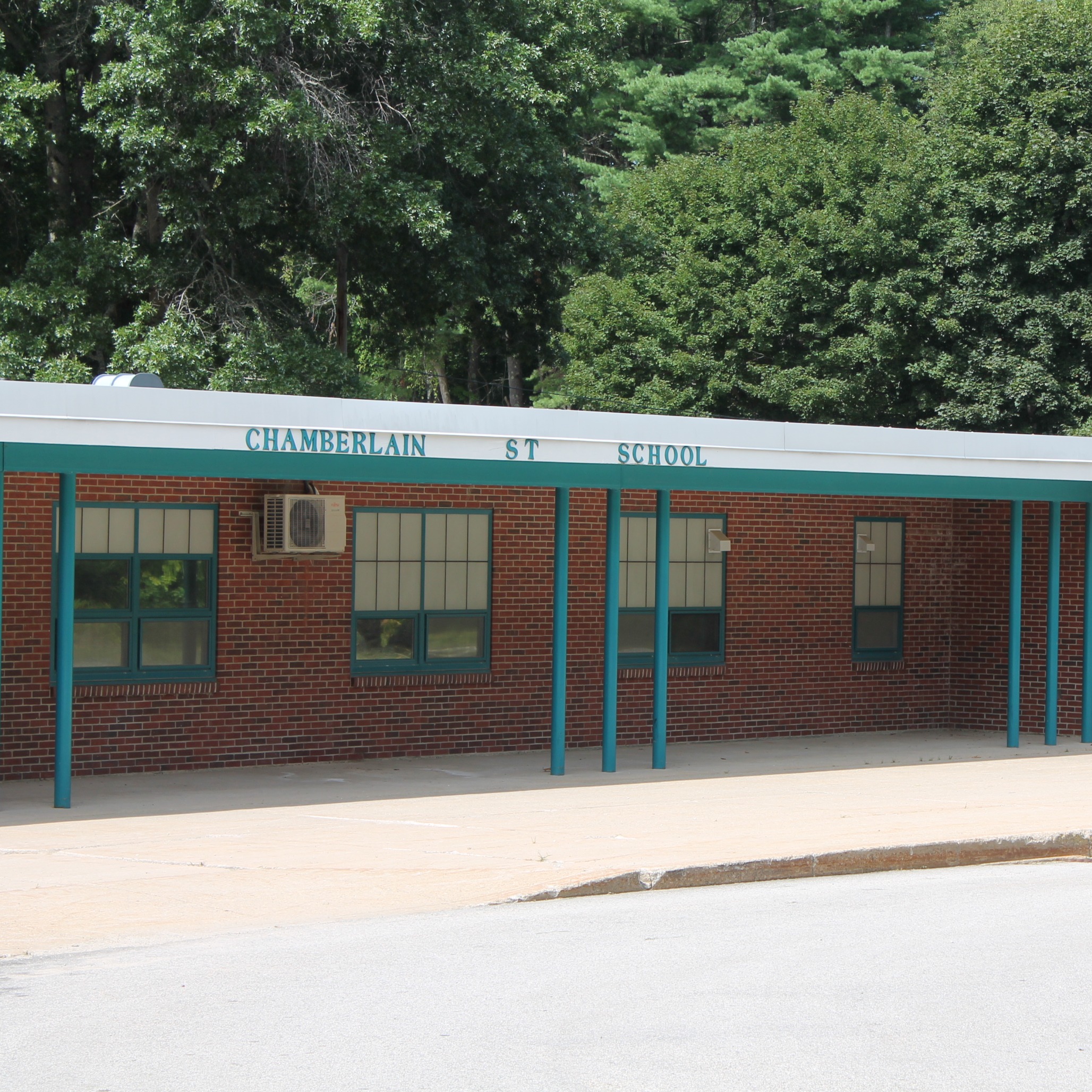 Brick building with white trim, labeled "Chamberlain 1st School," has green accents, large windows, and an air conditioner. Trees surround the area.