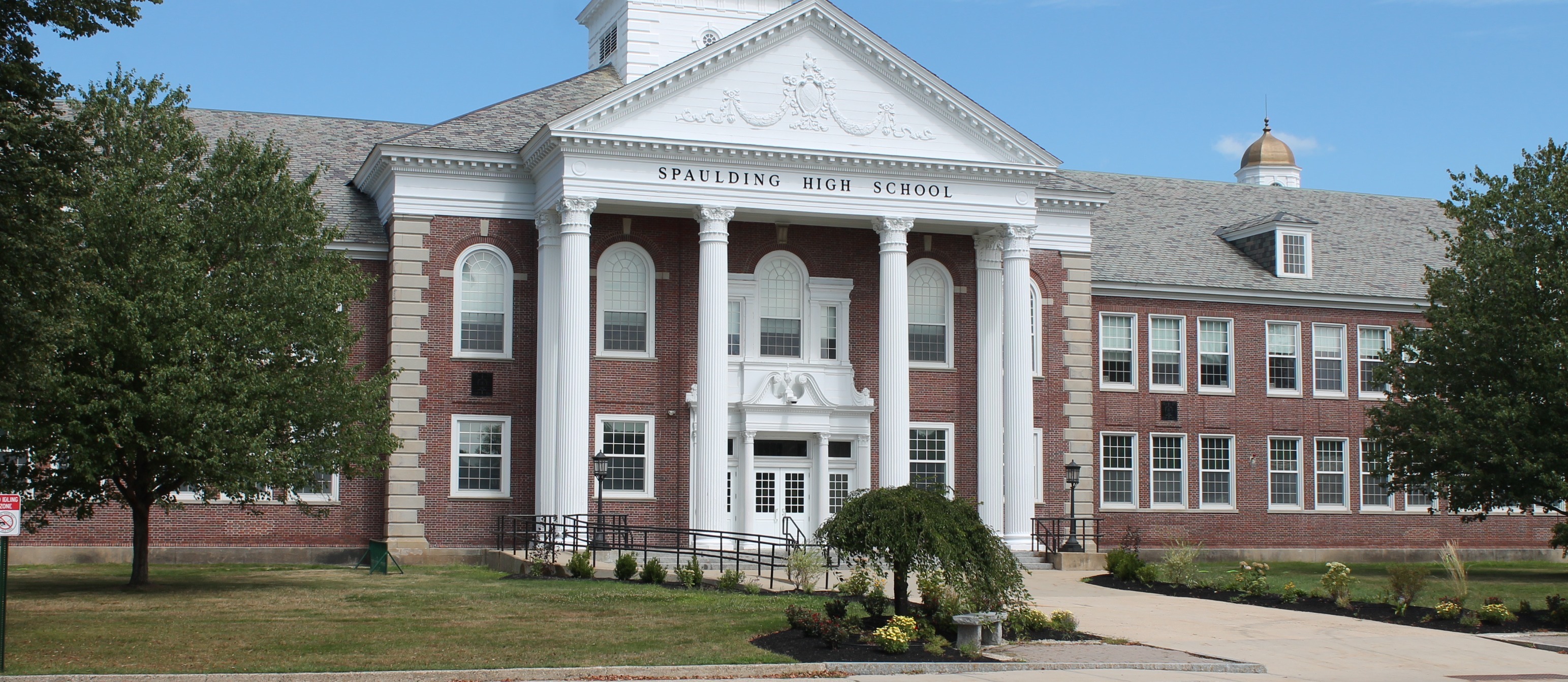 Spaulding High School. A brick building with a white tower and a clock, white pillars, and windows. A street and grassy area in front.