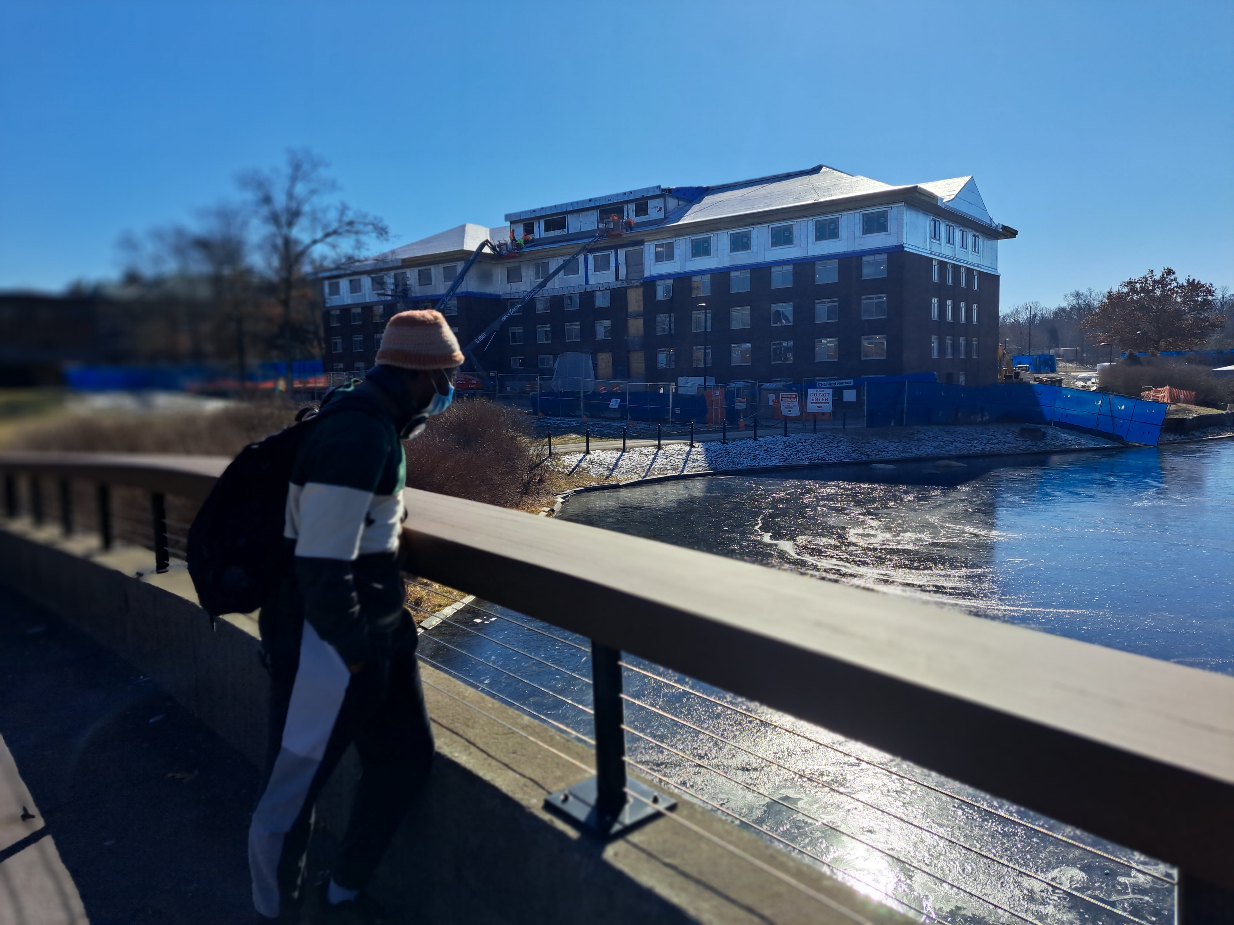 A student stands outside on a sunny day looking over a bridge at the water with a college in the background