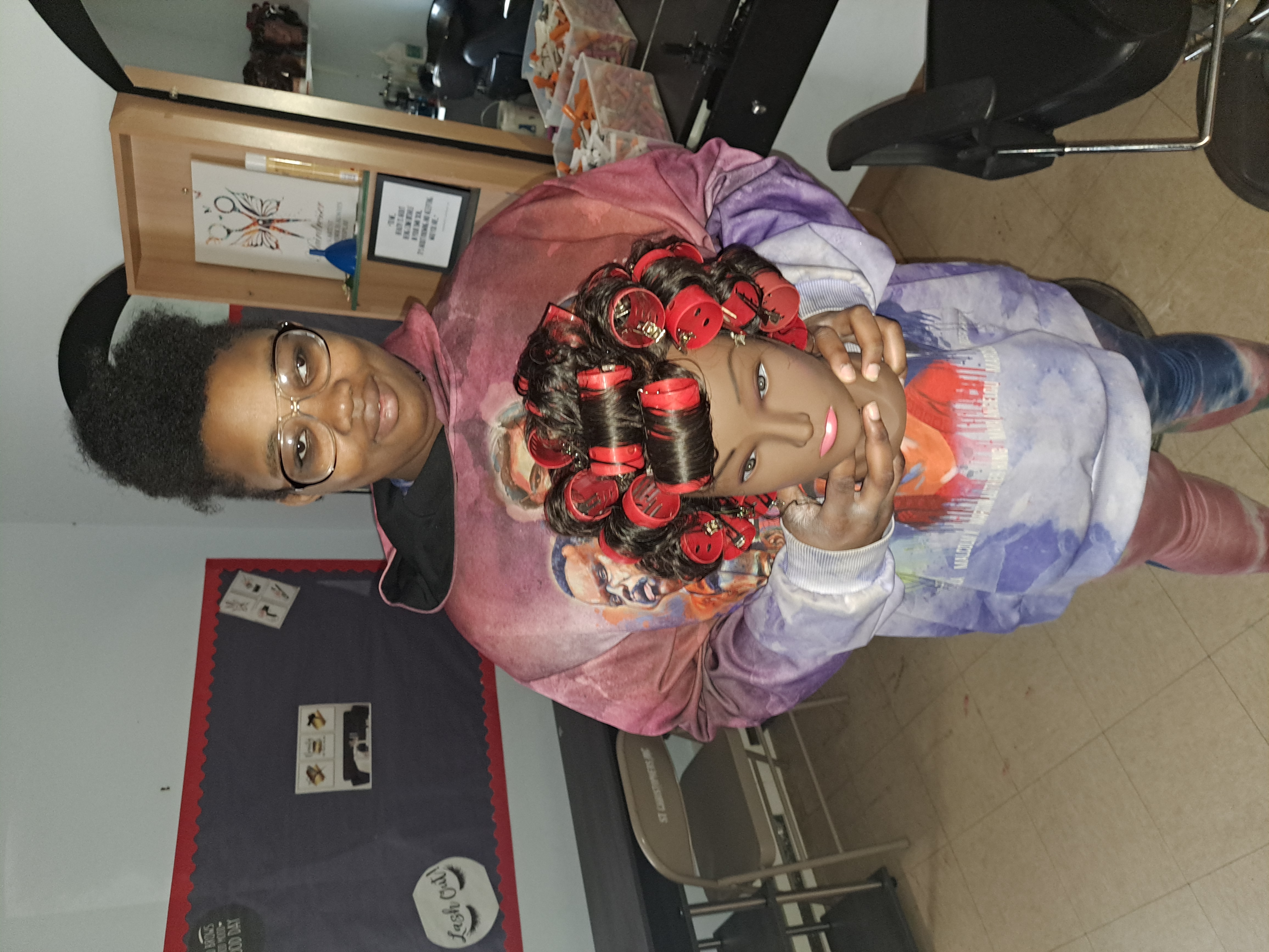 A student proudly holds a hair dressing mannequin head that she styled herself with many curlers