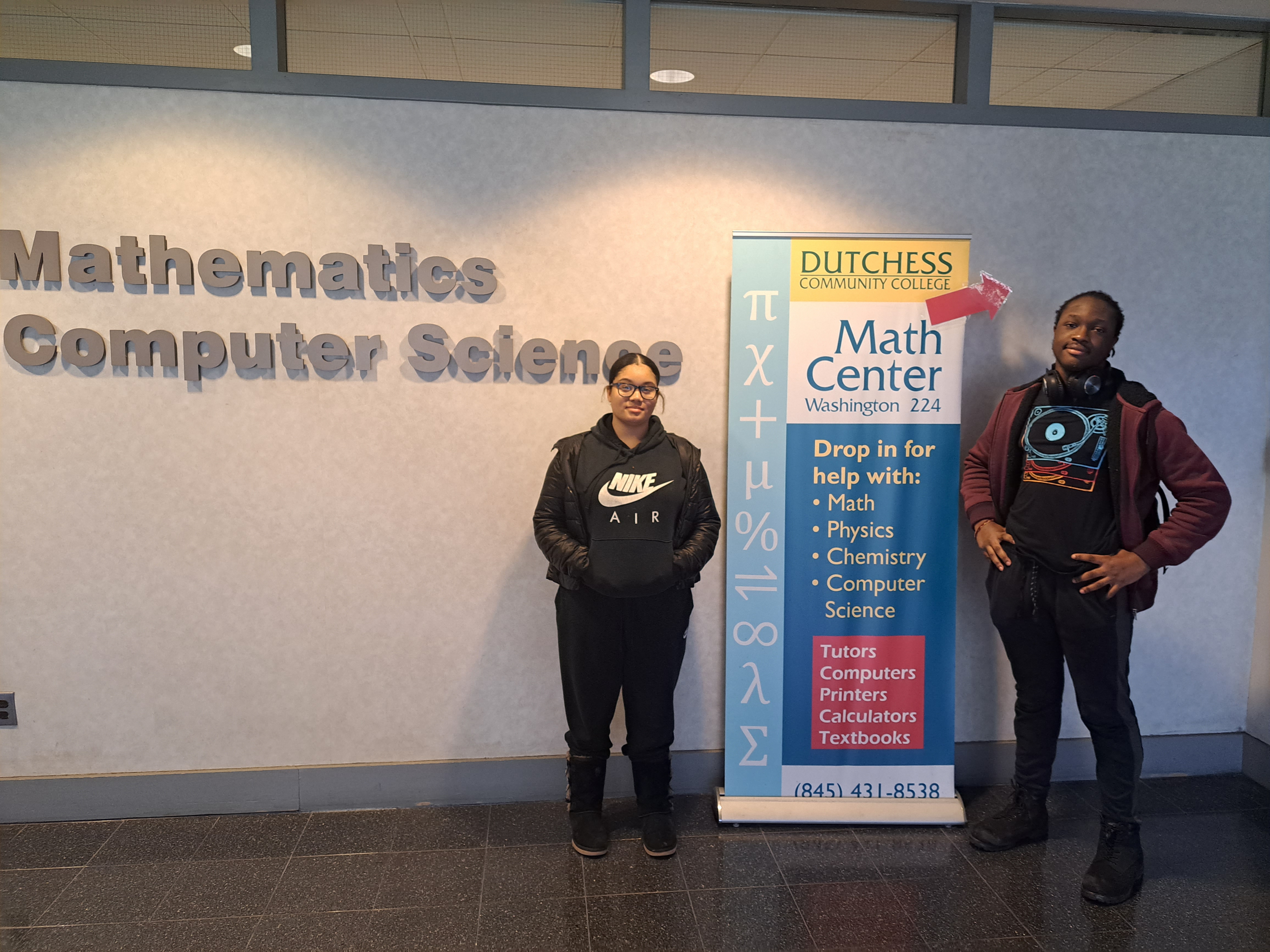 Two students pose inside dutchess community college at the mathematics computer science center by a sign that says "math center"