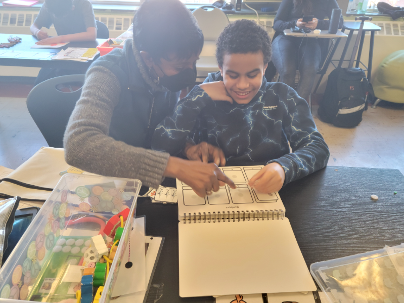 A classroom aide sits with a student at his desk helping him with his work