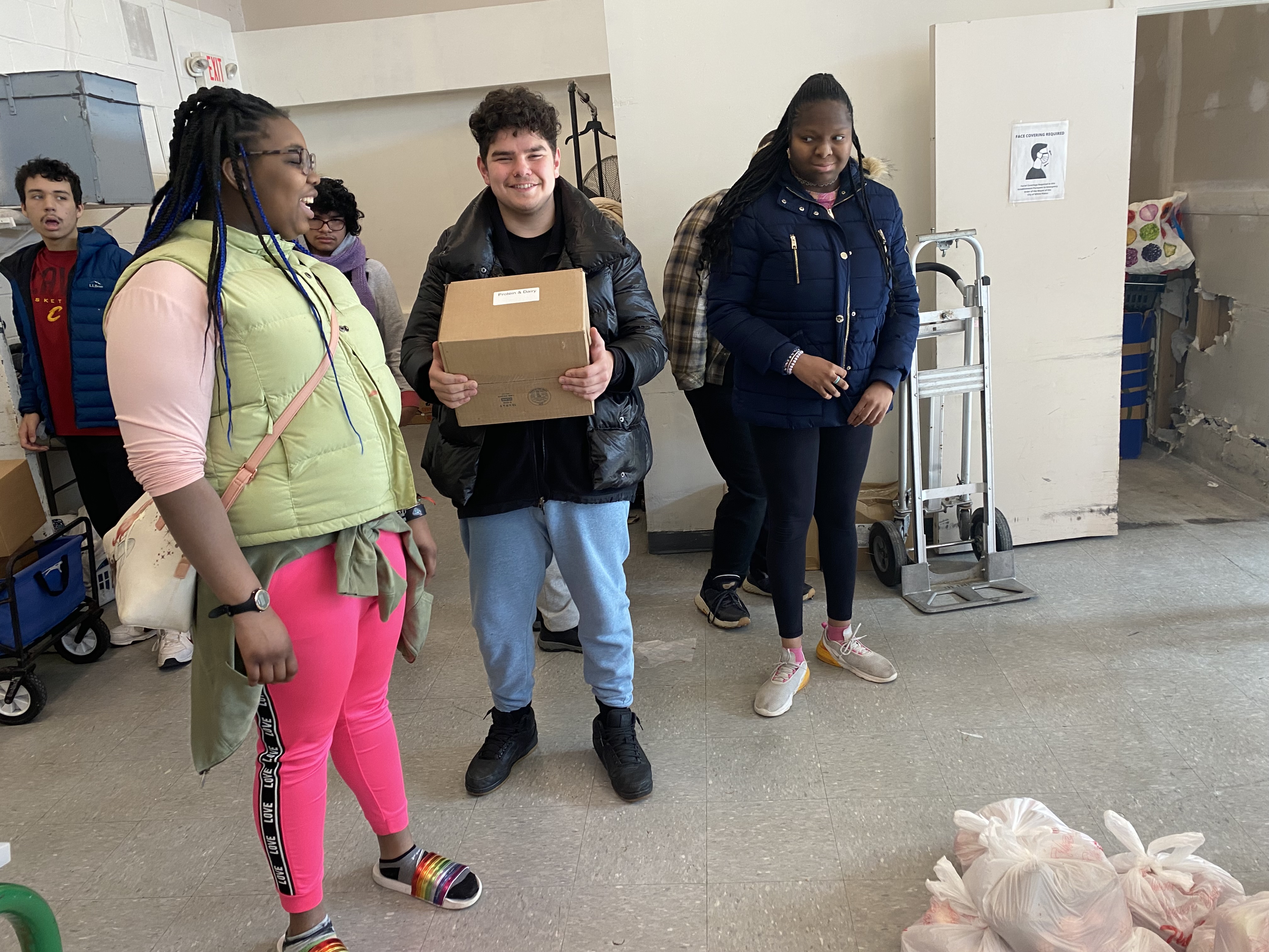 Students stand inside the food pantry smiling while one of them holds an open box of food