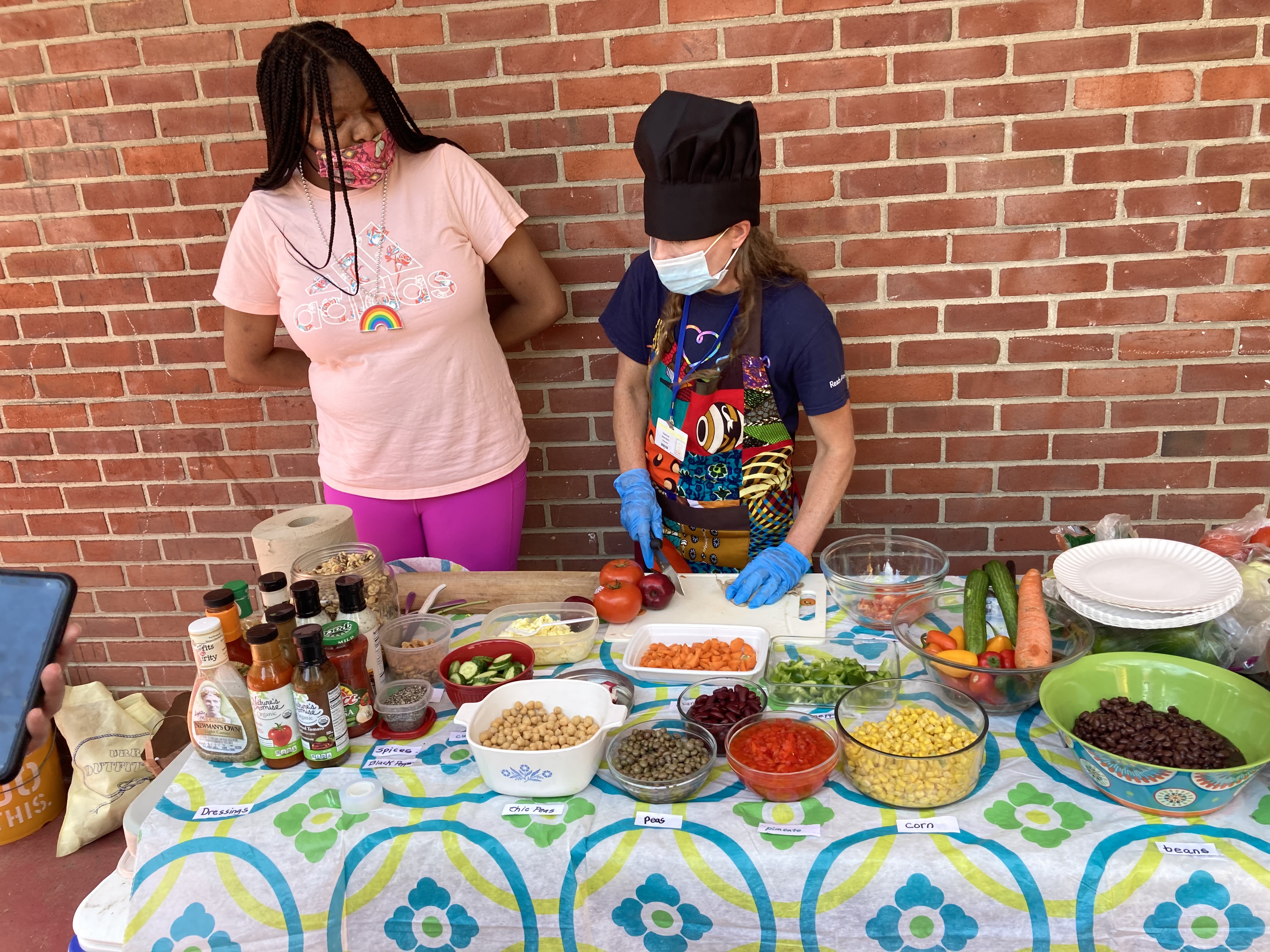A student stands next to Ms. Sheridan behind a table covered with food to help prepare it