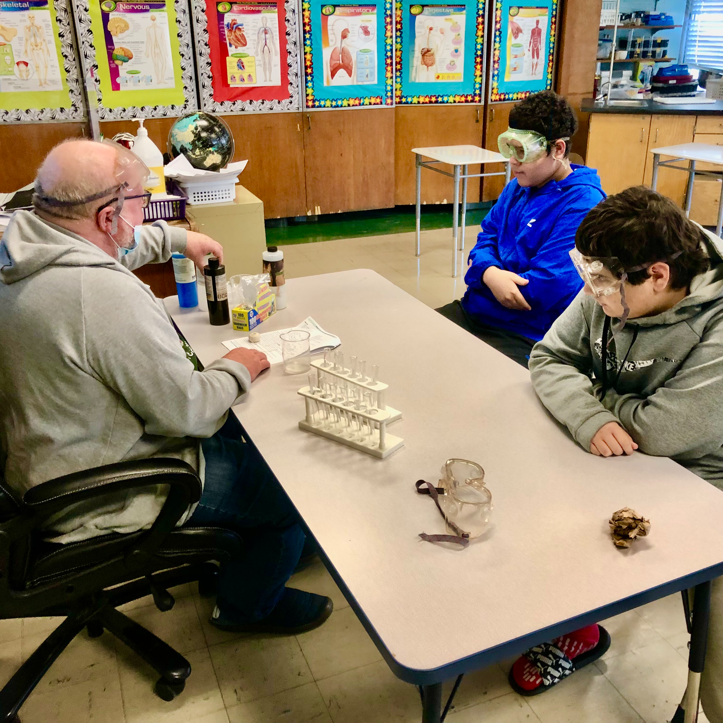 The science teacher sits at a table in the classroom with two students and everyone is wearing goggles to do an experiment